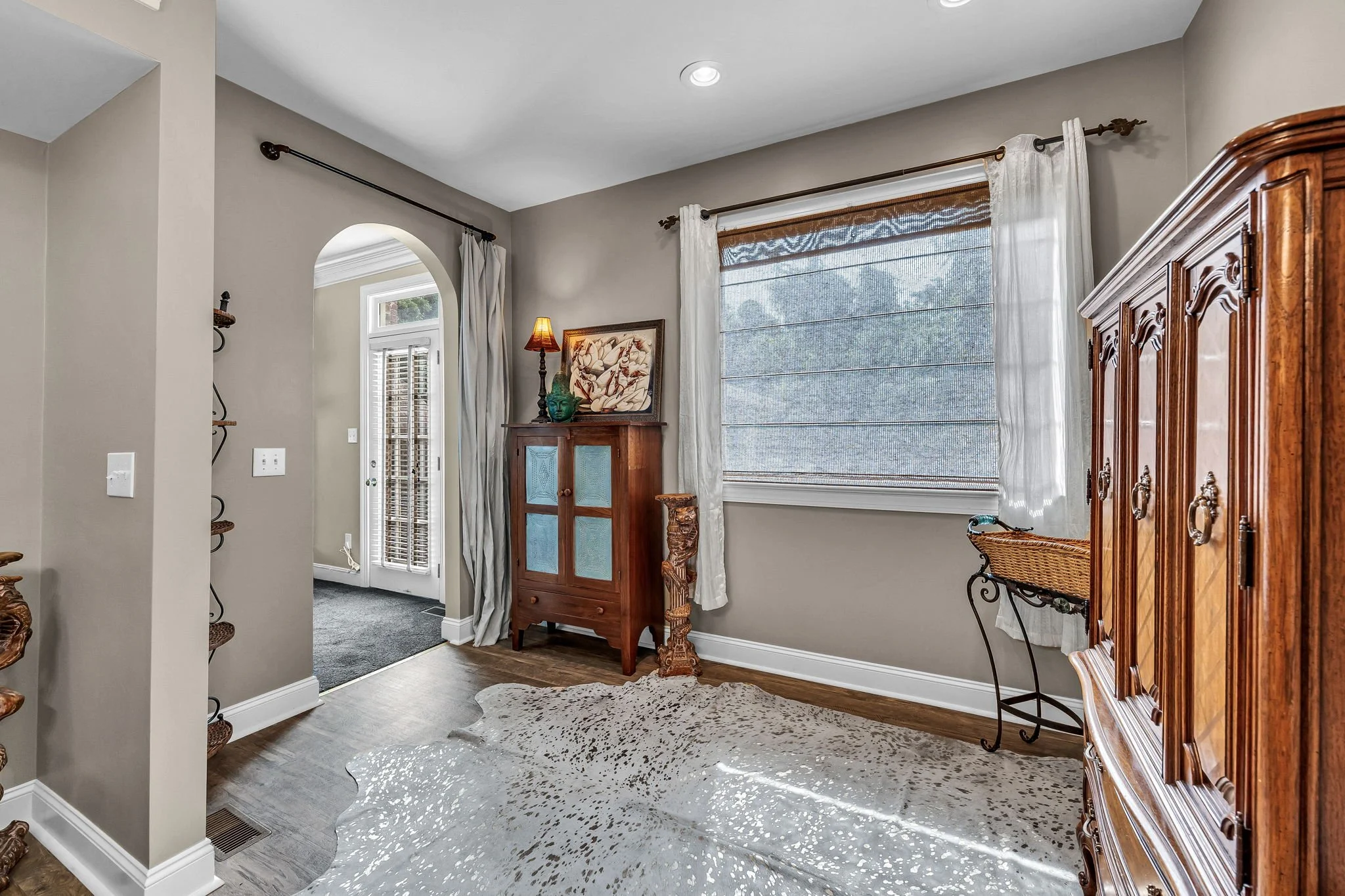 A cozy living room area with a large window covered by sheer curtains and wooden blinds, a wooden cabinet, a framed artwork, and a small table with a decorative lantern and a turquoise sculpture. An animal hide rug sits on the wooden floor, and a narrow hallway with a door leads outside.