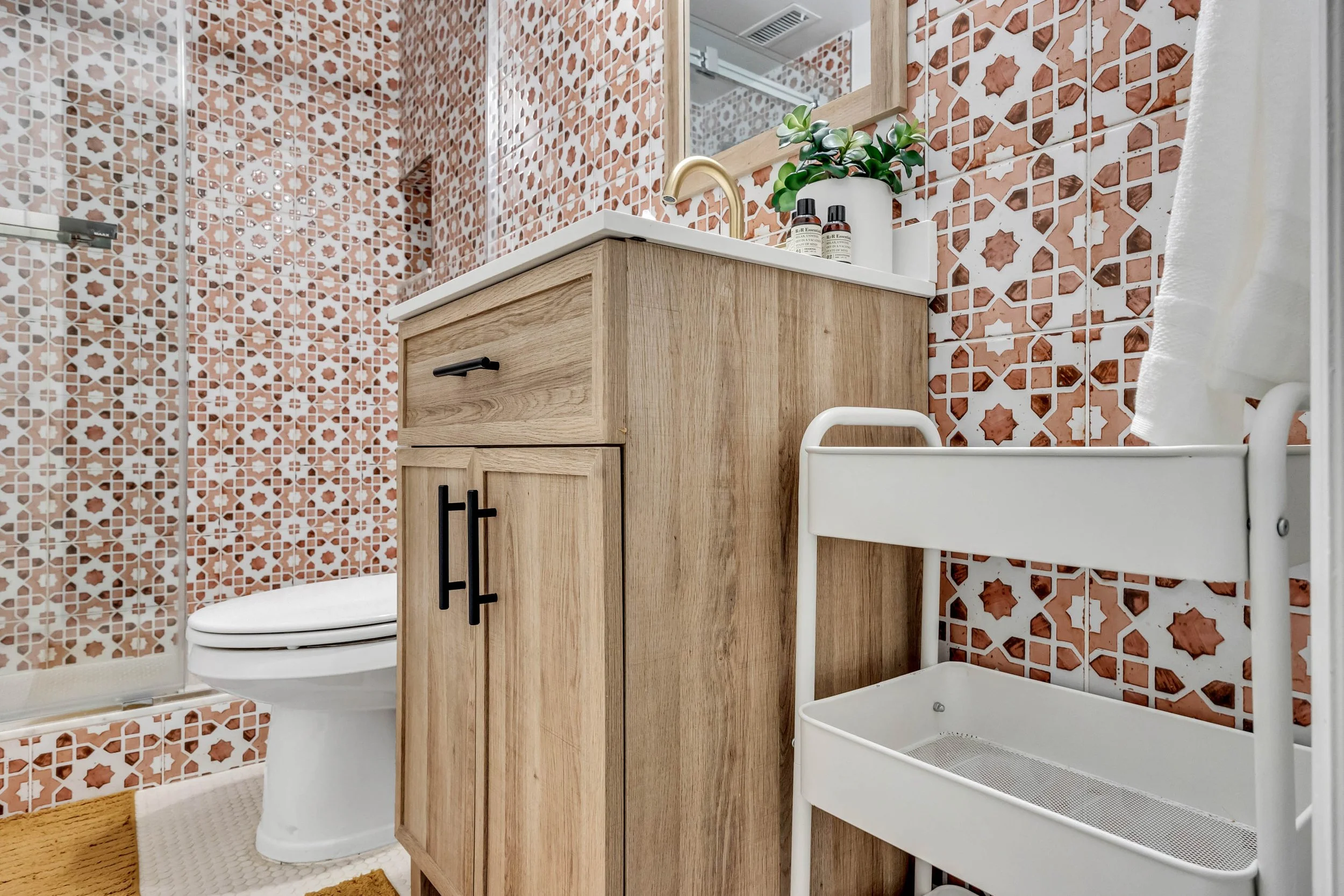 A bathroom with geometric patterned red and white wall tiles, a wooden vanity with a white countertop, a mirror above, a small potted plant, two toiletry bottles, a white towel hanging, and a white storage rack.