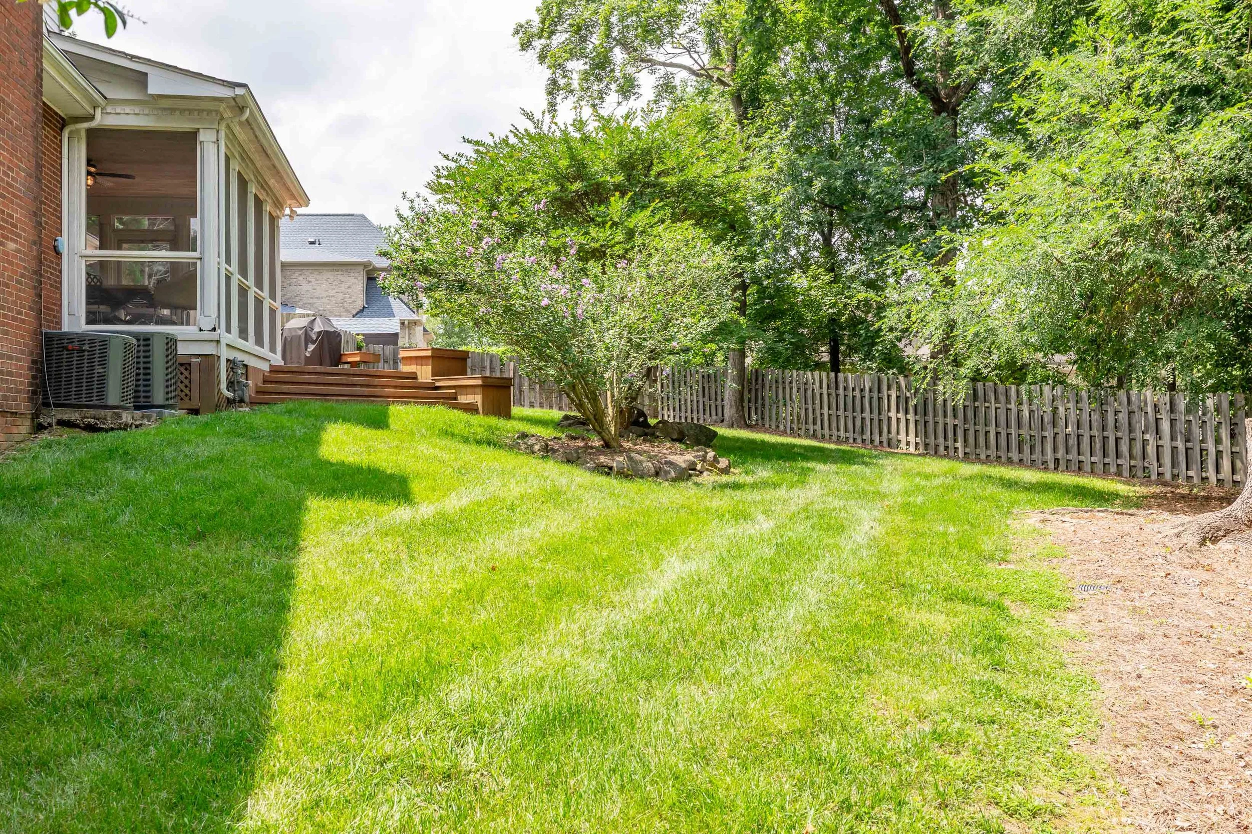 A backyard with a grassy lawn, trees, and a wooden fence. There is a house with a screened-in porch on the left, featuring an outdoor air conditioning unit below it. Wooden steps lead up to the porch, and there are neighboring houses in the background.