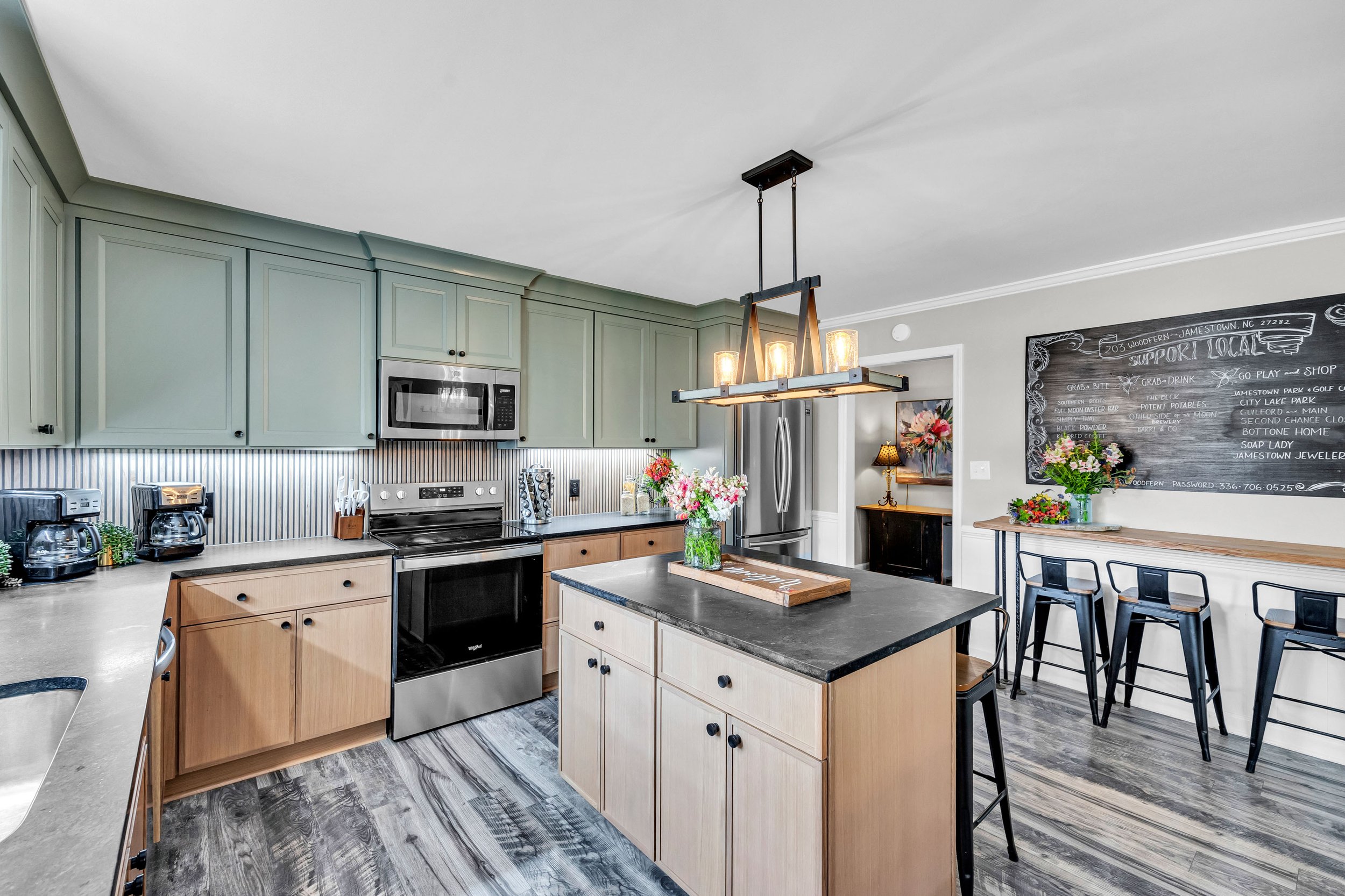 A modern kitchen with light green cabinets, a black countertop island with a vase of pink flowers, stainless steel appliances, a chalkboard wall with handwritten notes, and a black chandelier hanging from the ceiling.