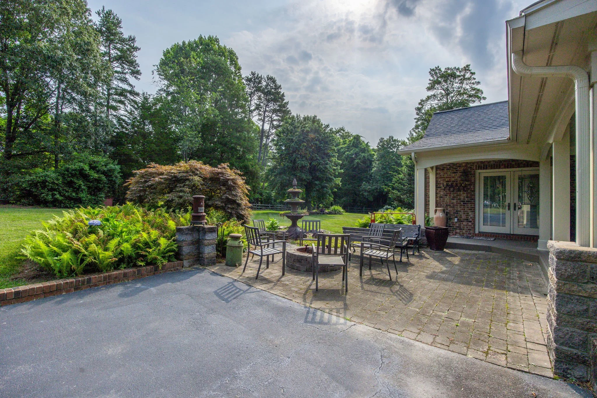 Residential backyard patio with outdoor furniture, a fountain, surrounded by lush green trees and plants, with a house on the right side under a partly cloudy sky.