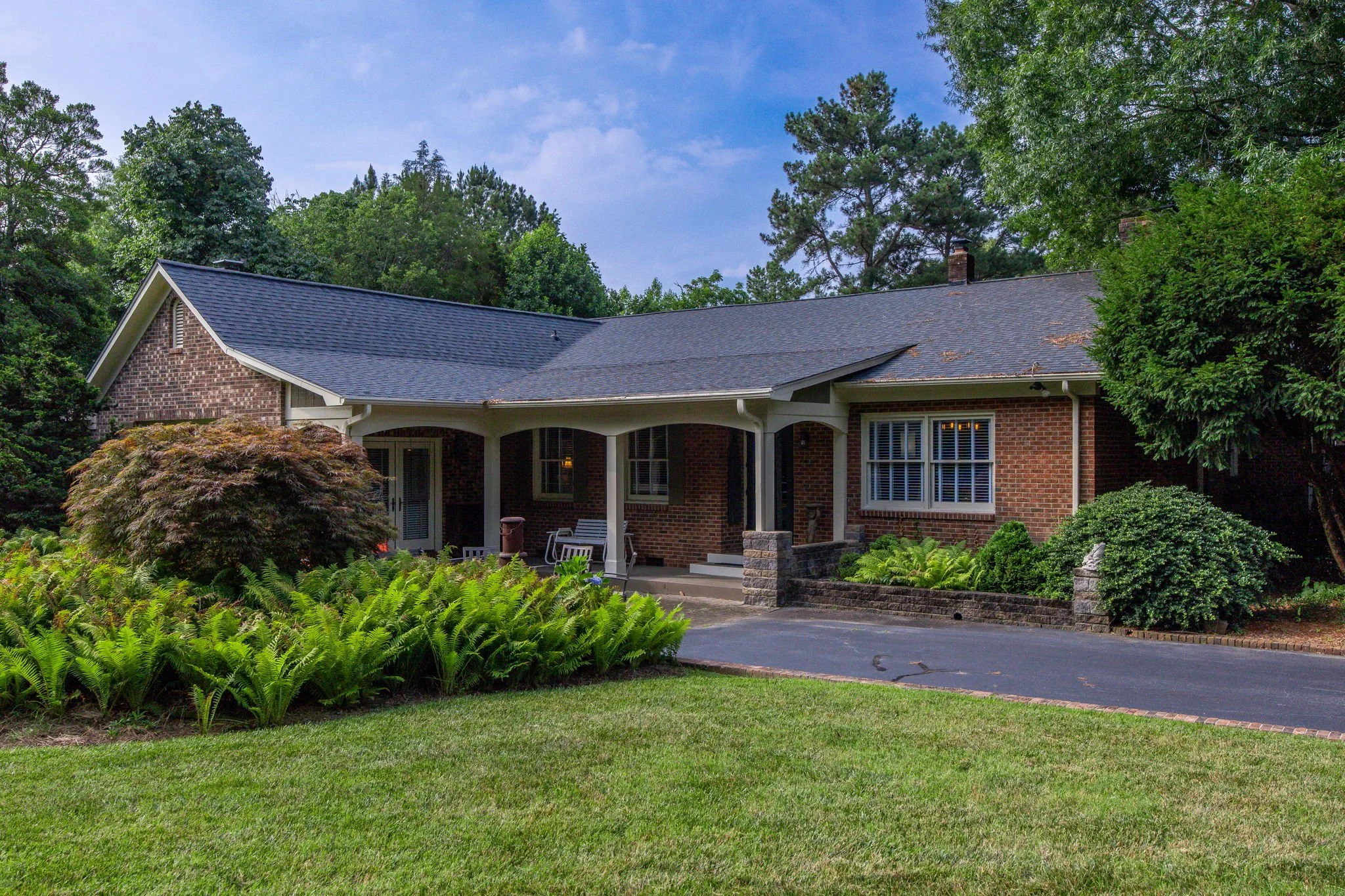A brick house with a dark gray shingle roof, surrounded by lush green trees and plants, with a grassy lawn in the foreground and a driveway leading to the entrance.