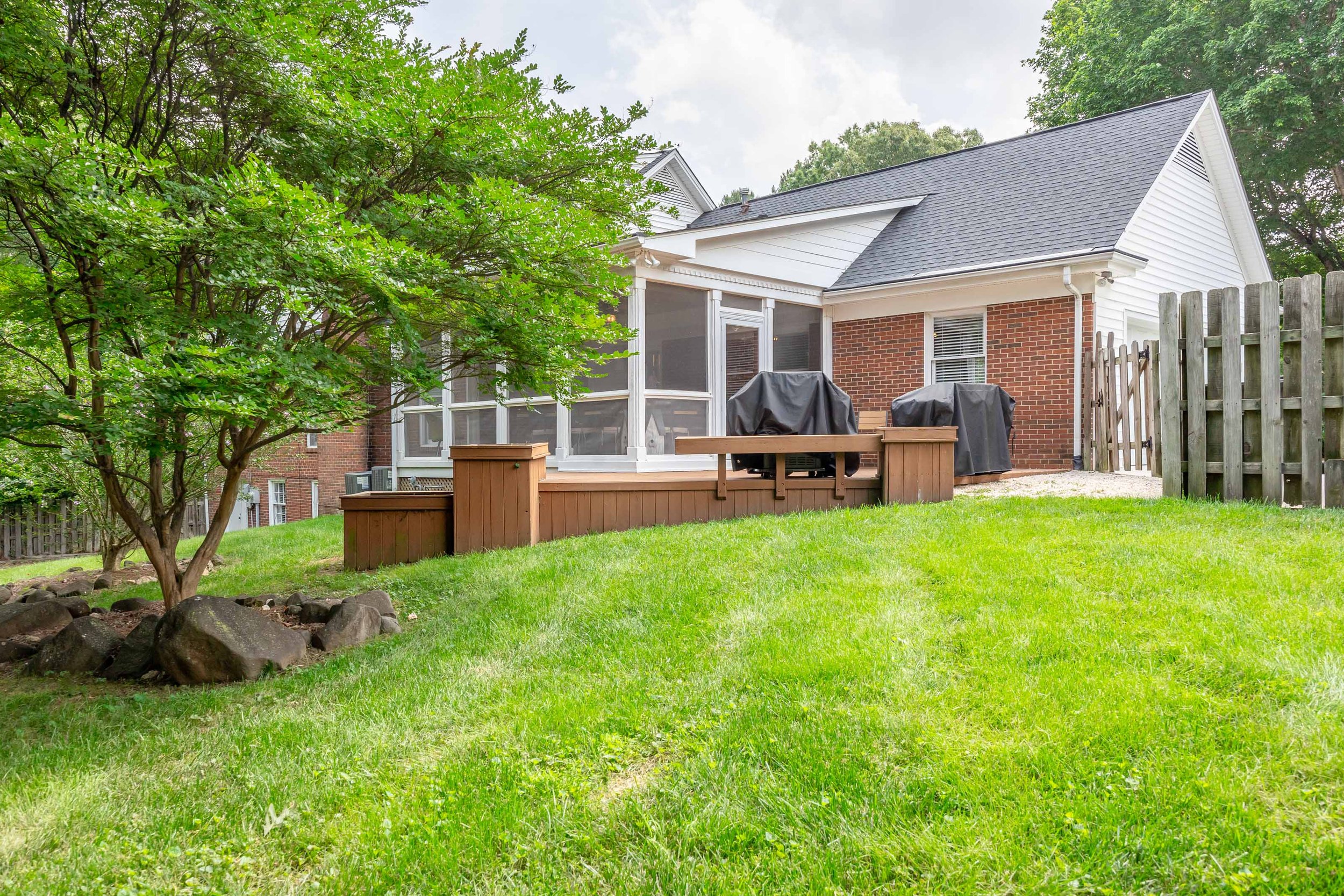 Backyard with green grass, a wooden deck with grills, a tree, a wooden fence, and the house with a shingled roof and white siding.