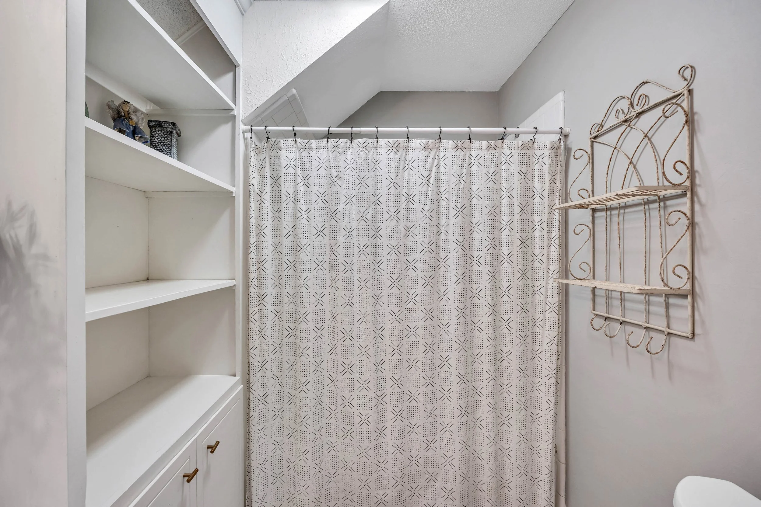 Interior of a small bathroom or closet with a white bookshelf on the left, a patterned shower curtain hanging in the center, and a decorative metal wall rack on the right. The ceiling has a sloped design.