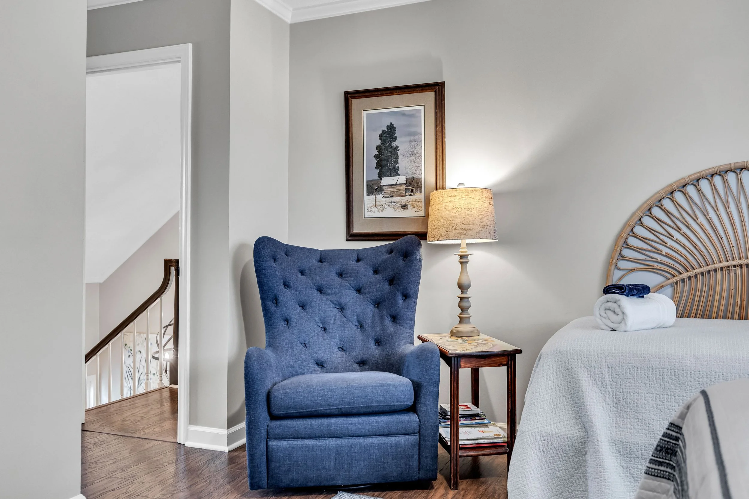 A bedroom corner featuring a blue upholstered armchair, a wooden side table with magazines, a beige table lamp, a bed with white bedding and rolled towels, a framed picture of a winter landscape on the wall, and part of a woven headboard. The room has hardwood floors and white walls.