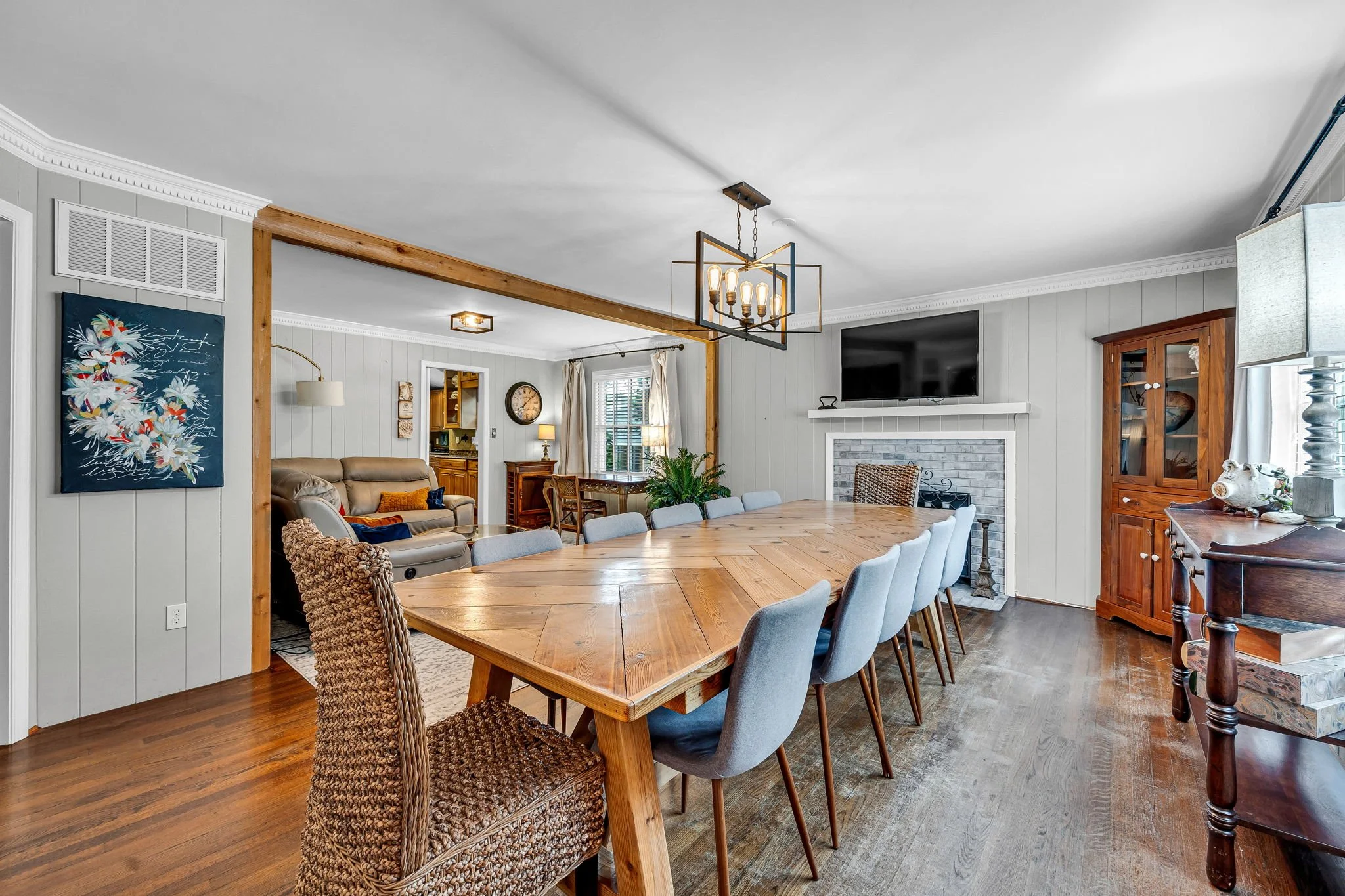 Spacious open-concept living and dining area with a large wooden dining table, light blue chairs, comfortable seating, a wall-mounted TV, fireplace, and decorative shelves, with natural light from windows.