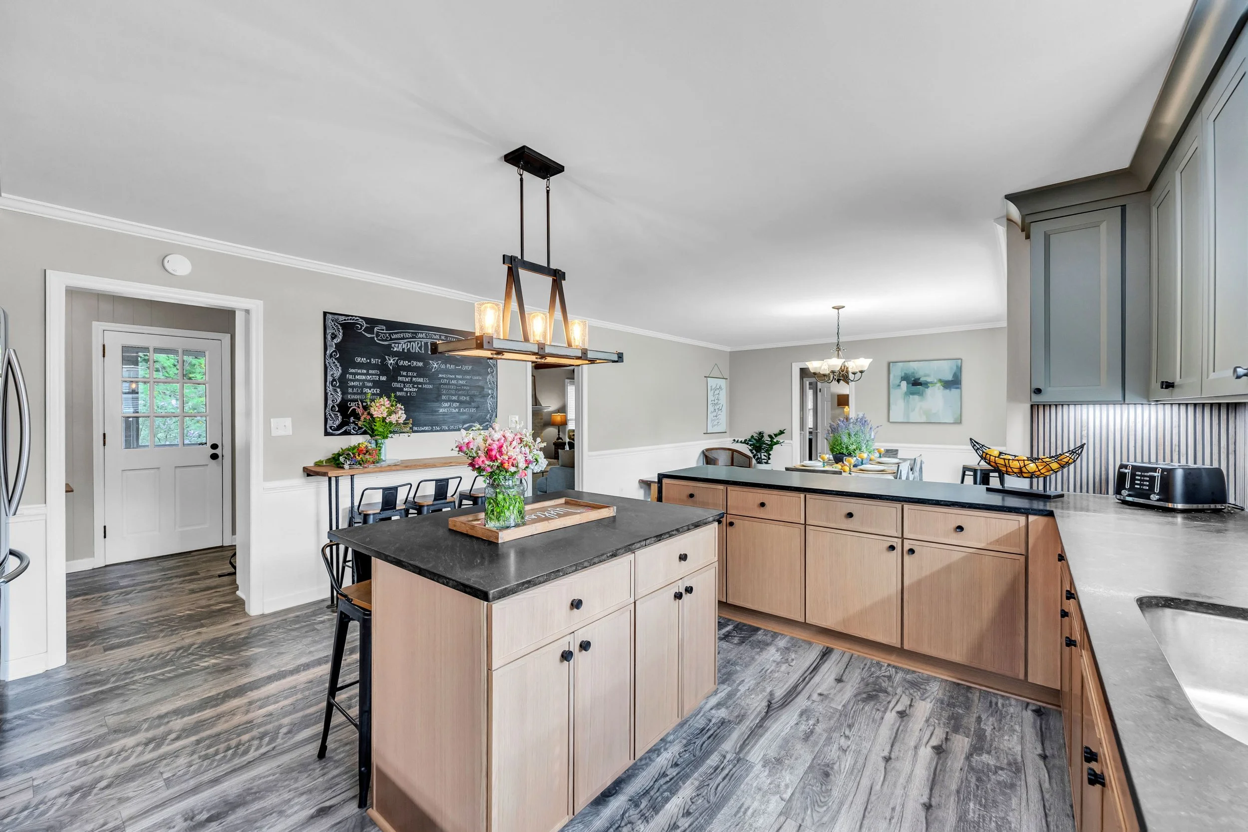 A modern, open-concept kitchen with light wood cabinets, a black countertop island, and a dining area with a table set for meals, decorated with flowers and lemons. There are pendant lights hanging above the island and natural light coming through the windows.