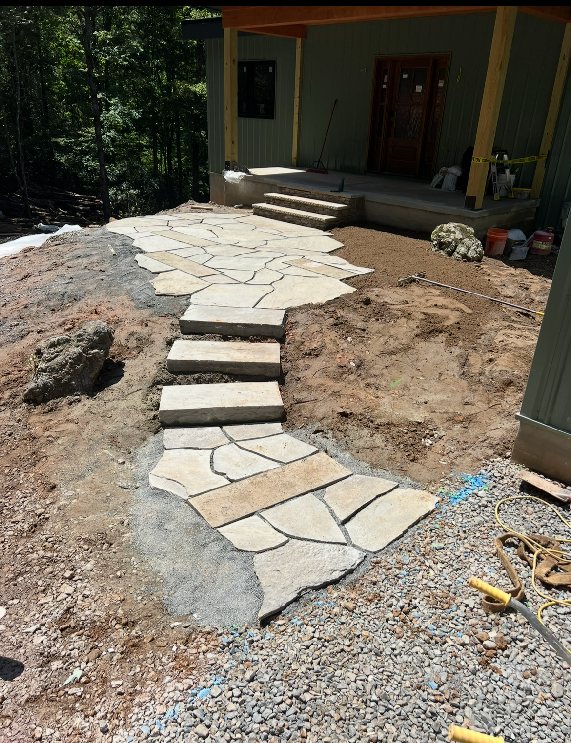 A paved stone pathway in construction leading up to a house with a porch. The pathway has irregularly shaped stones and wooden steps, with dirt and gravel surrounding it. In the background, trees are visible.