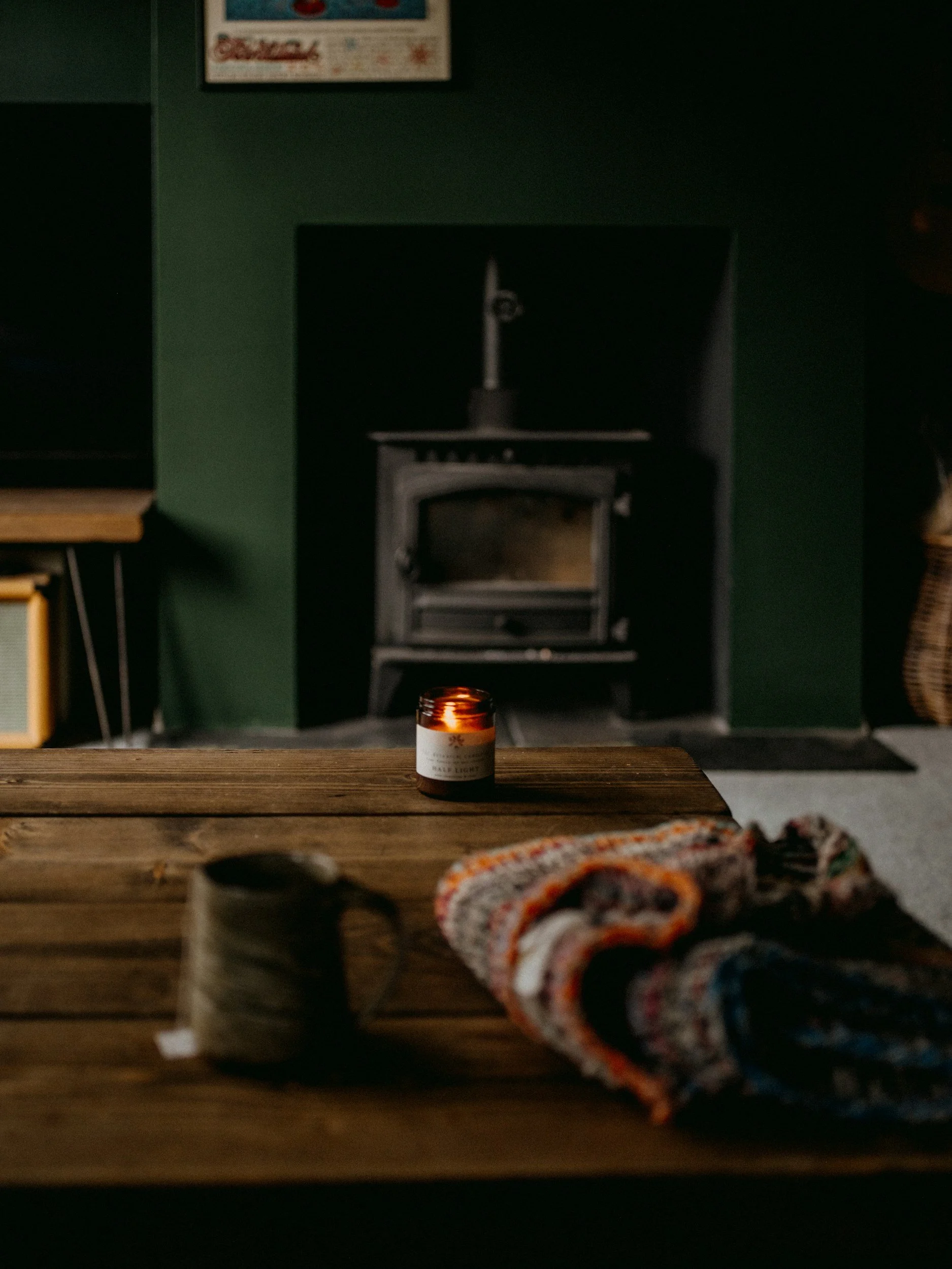 A candle on a wooden table, with a black stove and dark green wall in the background, a blanket on the table, and a basket on the wall to the right.