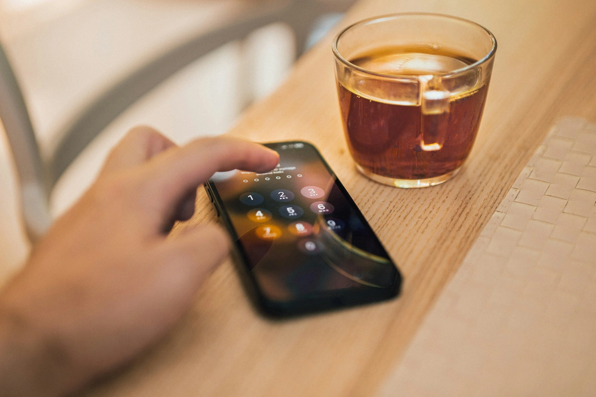 A hand holding a smartphone with a numeric passcode screen visible, next to a glass of iced tea on a wooden surface.