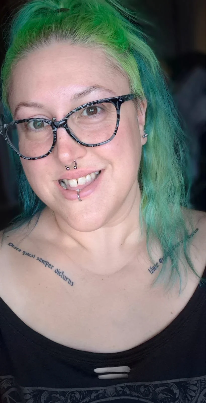 A woman with green and blue hair, wearing patterned glasses, with multiple facial piercings, and tattoos on her chest.