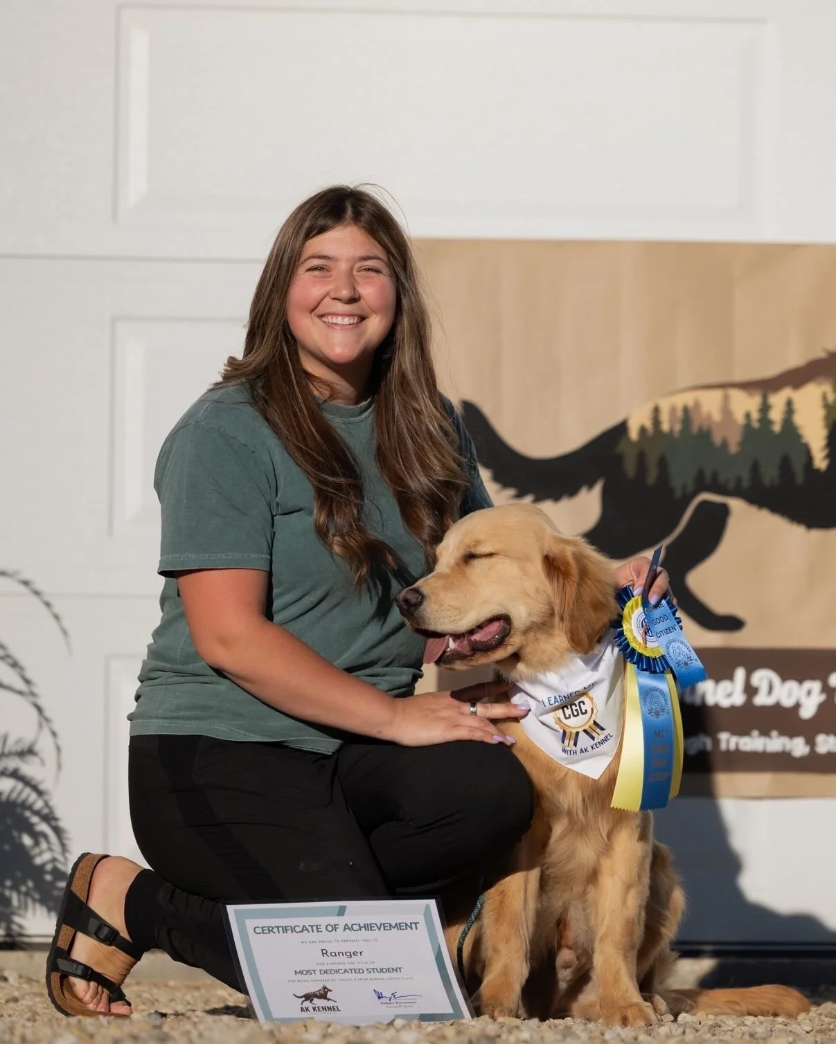 A girl kneeling next to a golden retriever wearing a ribbon, holding a certificate of achievement in front of a backdrop with a wildfire logo and trees.
