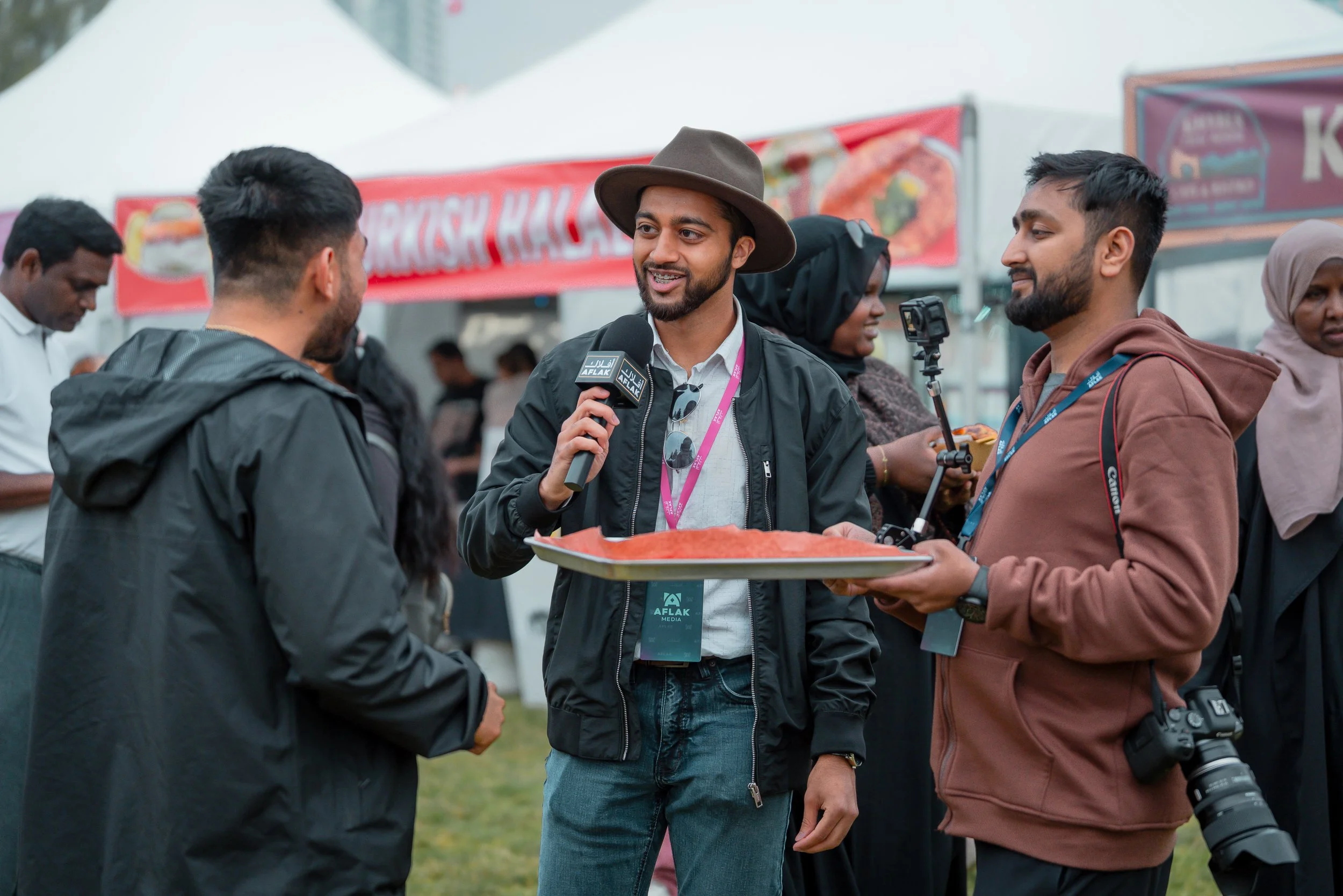 A man wearing a brown hat and black jacket is speaking into a microphone labeled 'AFLAK Media' while holding a tray of food at an outdoor event. Two other men are listening, one with a camera and another holding a camera, with tents and banners in the background.