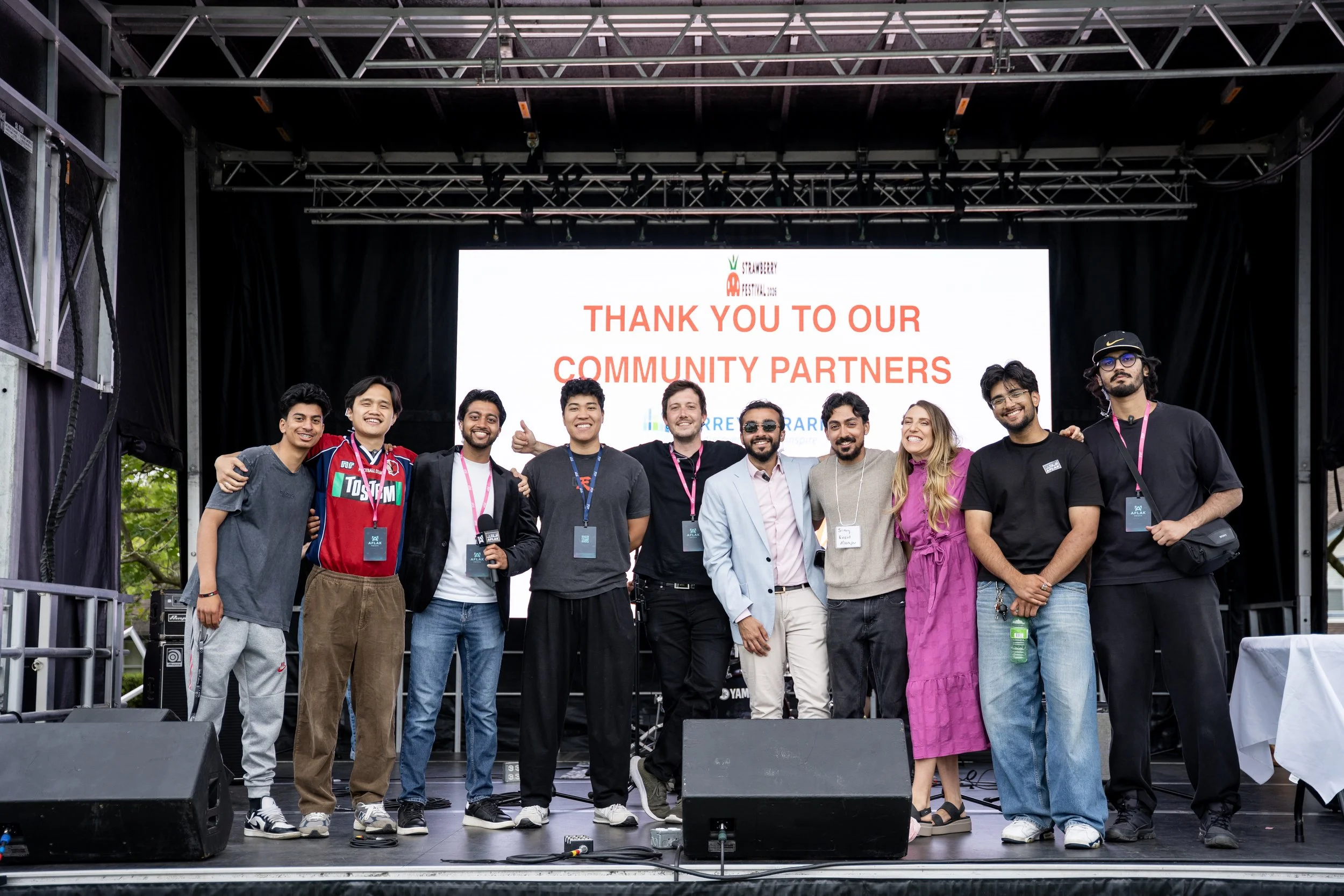 A group of eleven diverse people standing on an outdoor stage, smiling and posing for a photo with a large screen behind them displaying a message of gratitude to community partners. The stage has black curtains and metal scaffolding.