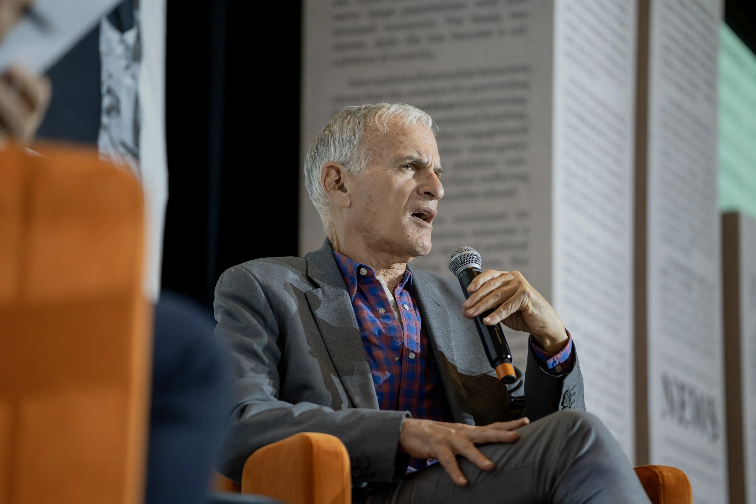An elderly man with gray hair speaking into a microphone during a panel discussion or interview, sitting on an orange chair, wearing a gray blazer and a plaid shirt.