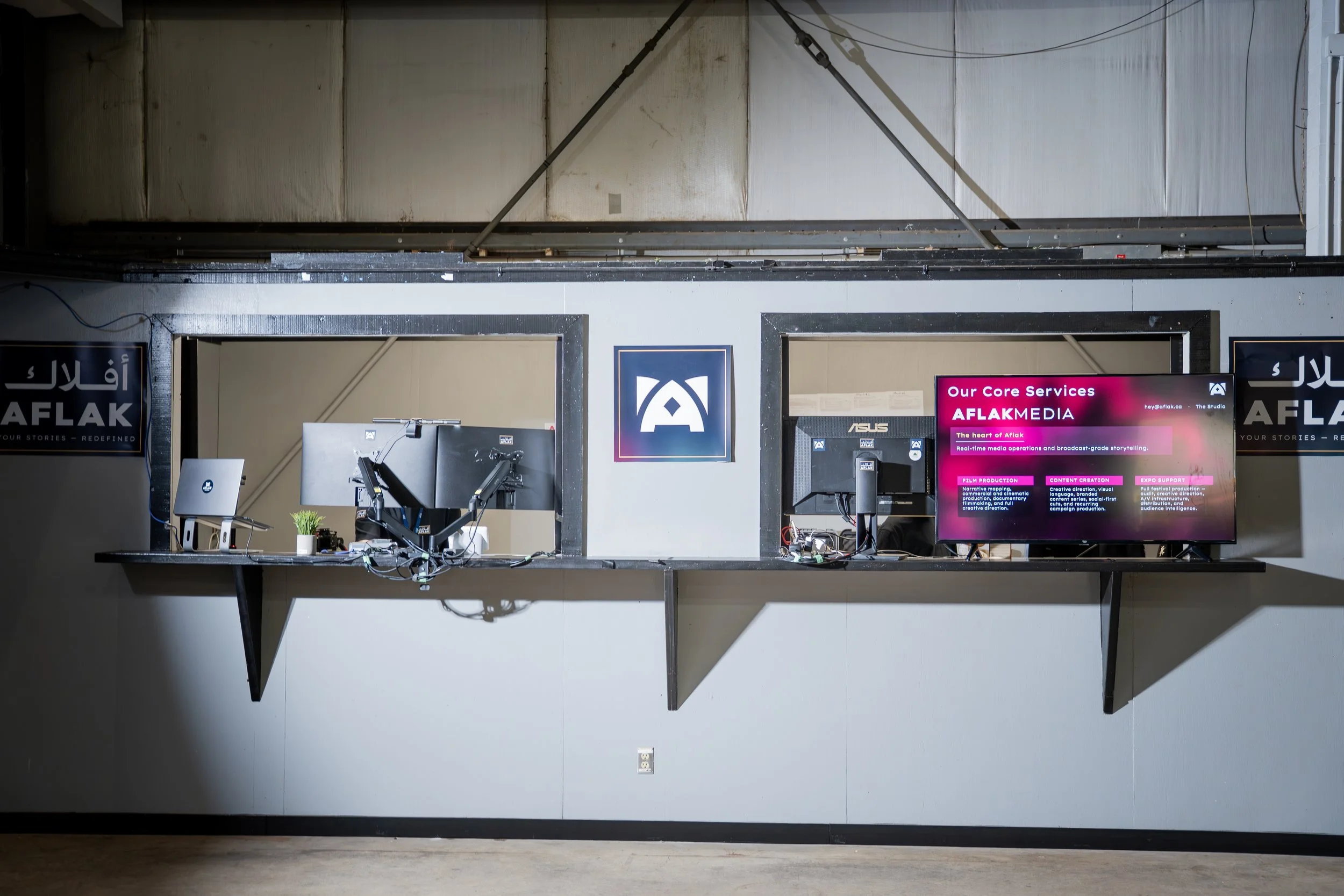 A workspace with computer monitors, keyboards, and a desk, situated behind a large opening in the wall. The background shows an unfinished ceiling with pipes and wires, and signs for AF Lean media.