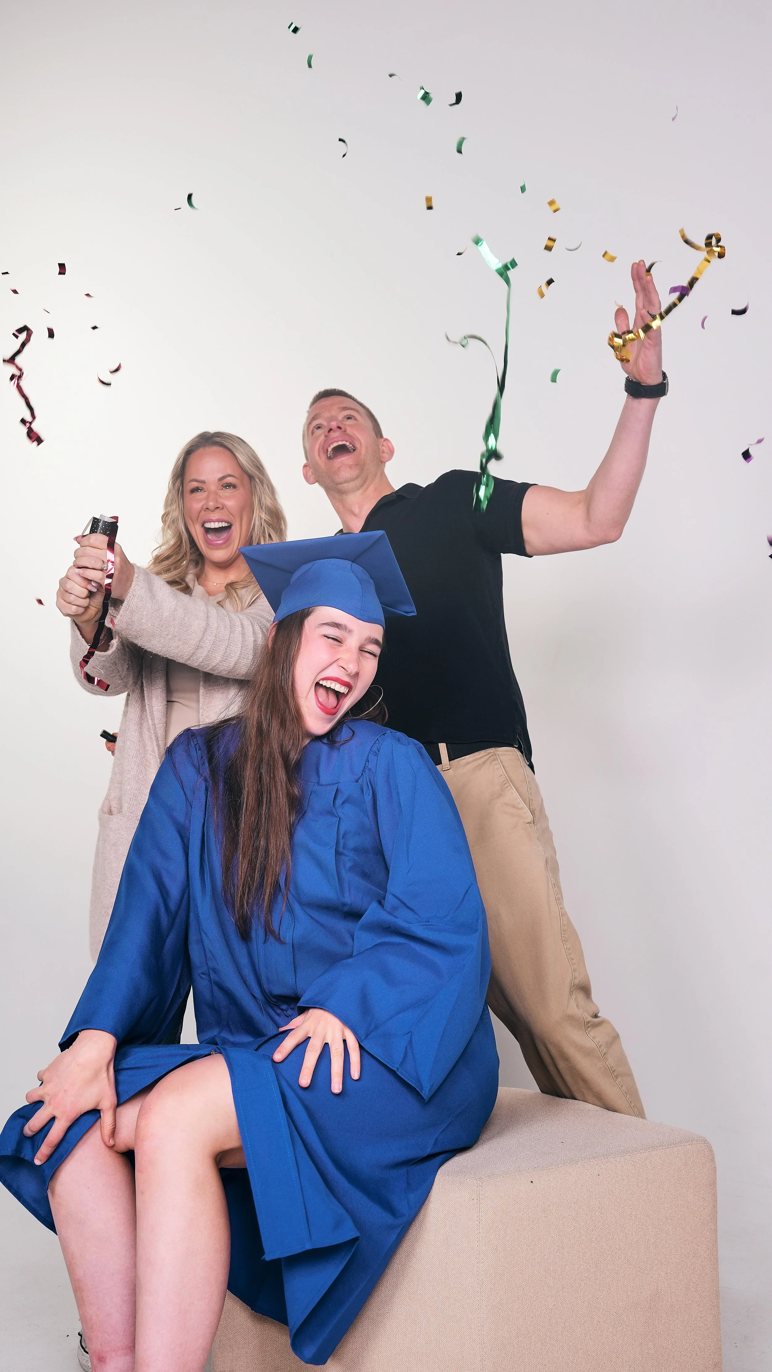 A young woman in a graduation cap and gown sitting on a couch, surrounded by celebrating adults, with confetti in the air.