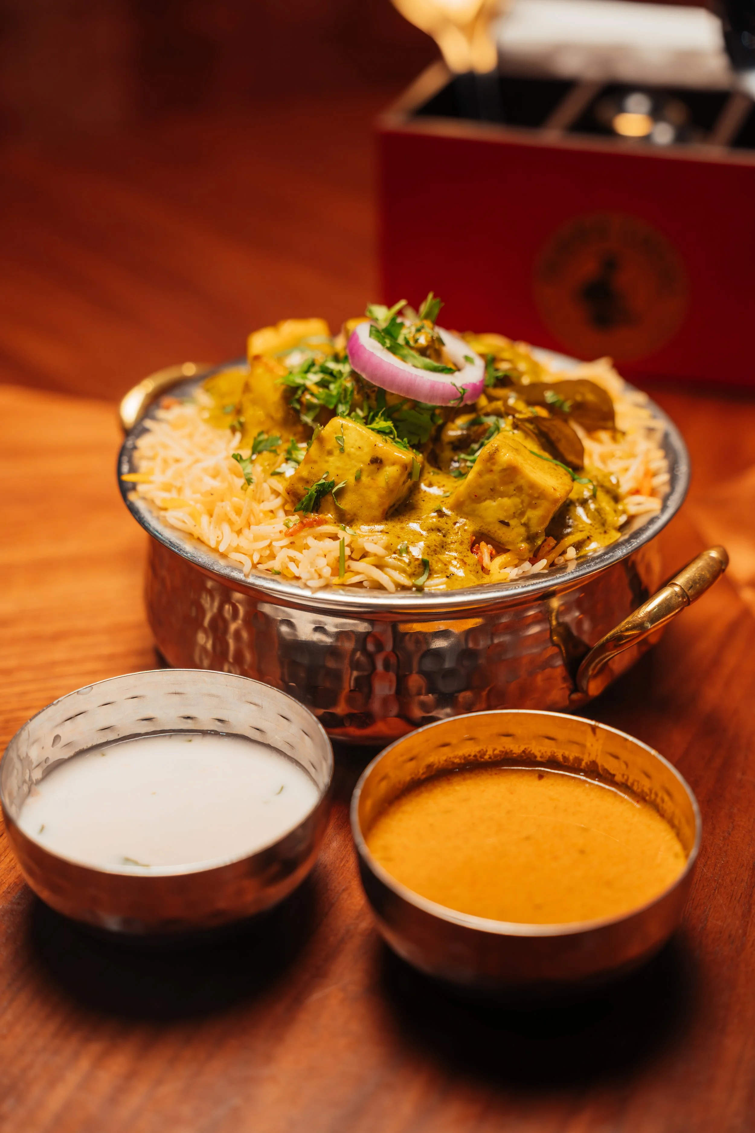 Close-up of Indian curry and rice served in a metal bowl with two small copper cups of yogurt and sauce placed in front on a wooden table.