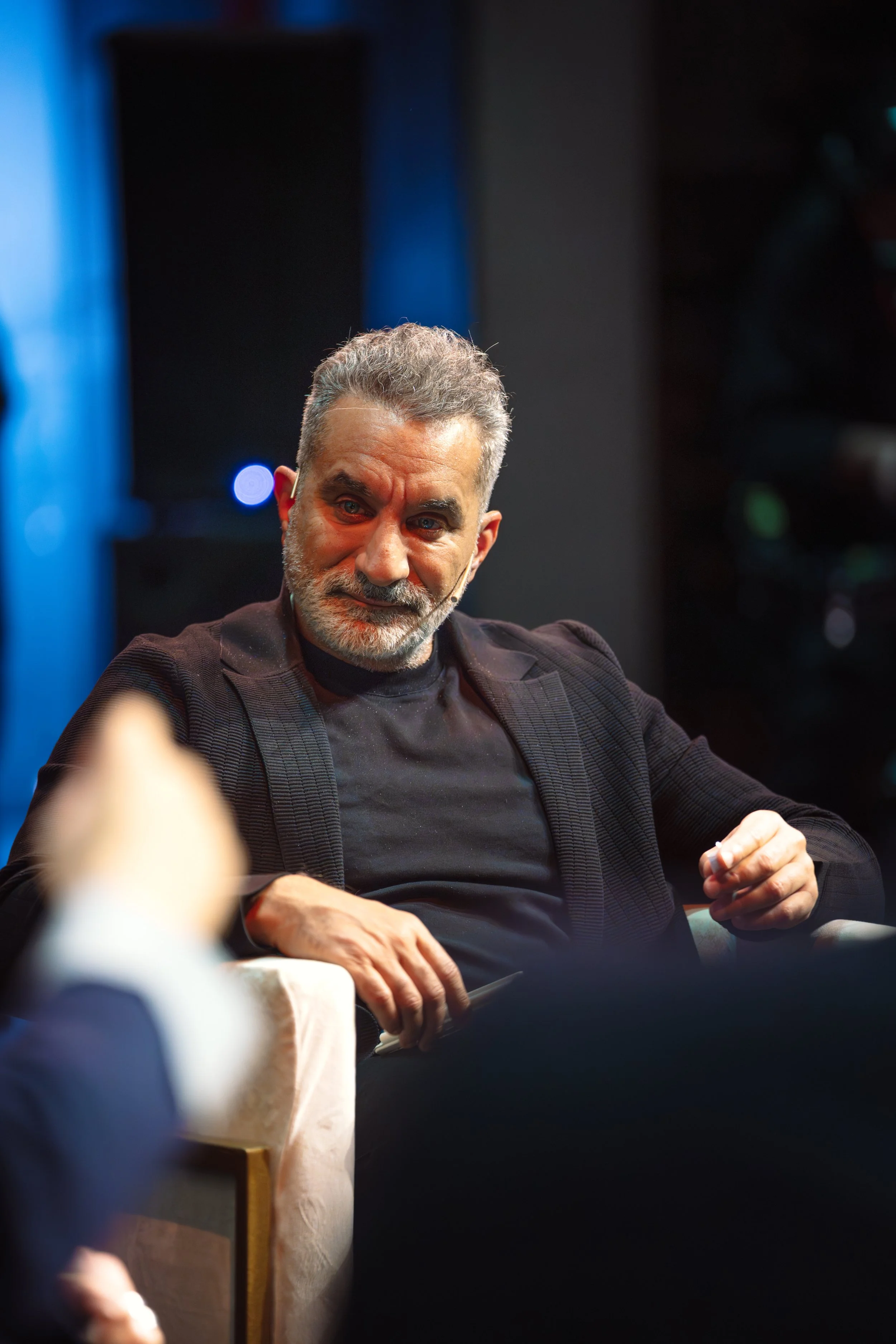 Portrait of a middle-aged man with gray hair and beard, wearing a dark checked blazer over a black shirt, smiling while sitting at an event with a dark background and a blue-lit area on the left.