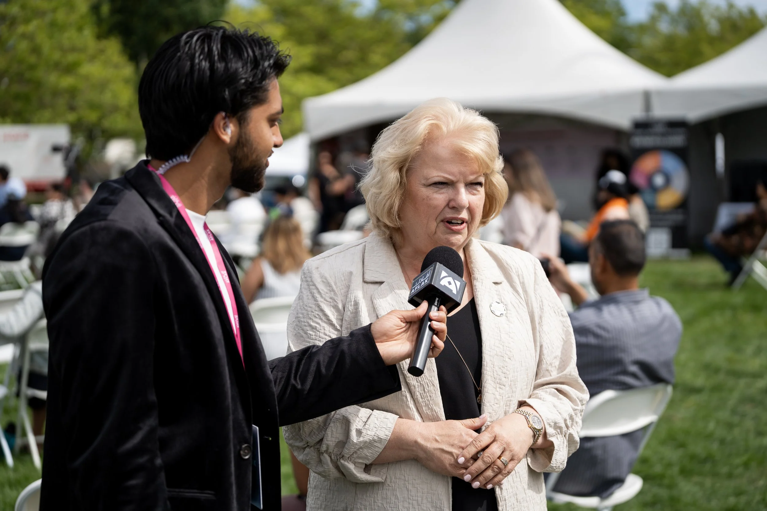 A woman with blonde hair being interviewed by a man holding a microphone at an outdoor event with tents and people in the background.
