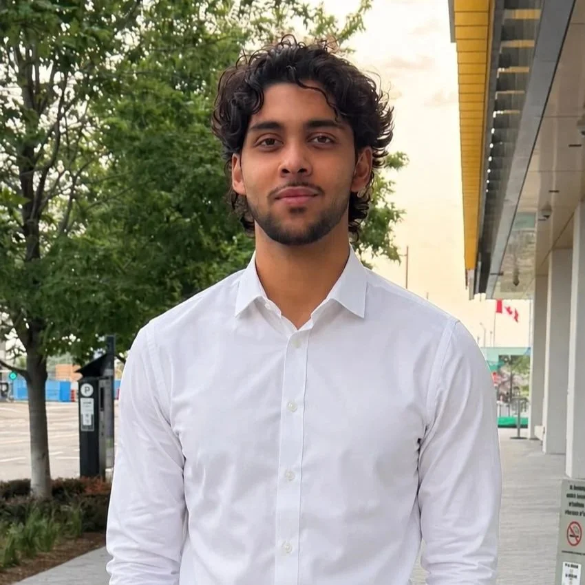 A young man with curly dark hair and a beard, wearing a white dress shirt, standing outdoors near some trees and buildings.