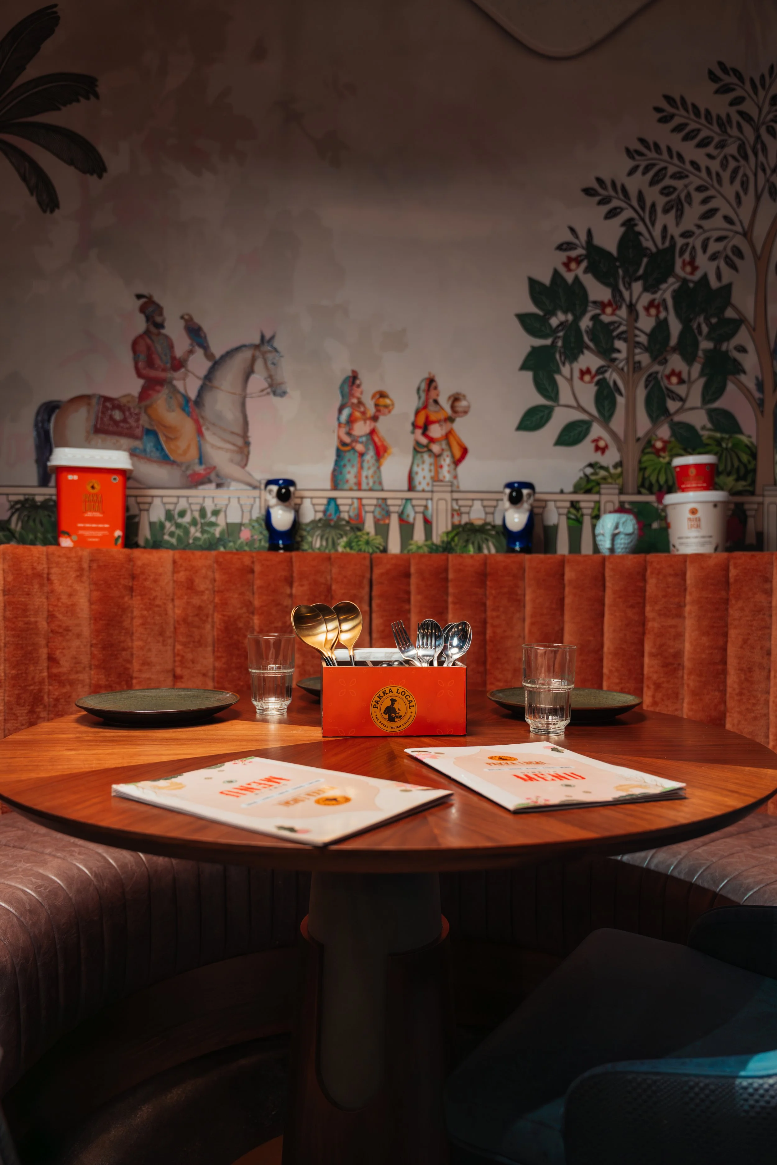 A restaurant table setting with menu, glasses, and cutlery, decorated with wall art depicting traditional Indian dancers, a person on horseback, a large tree, and potted plants in the background.