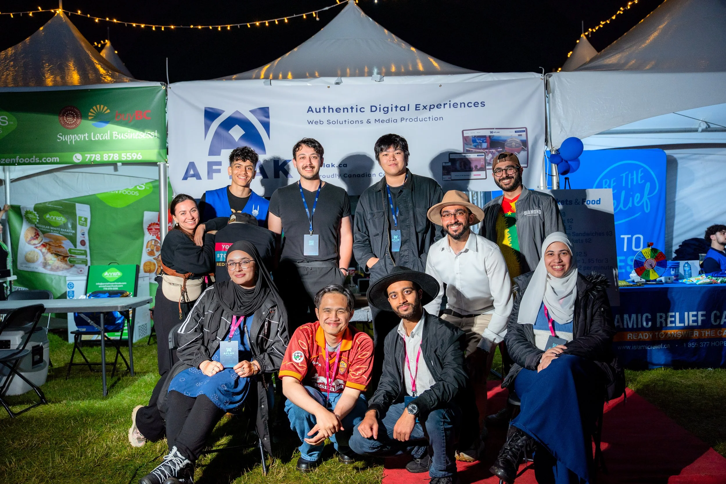 Group of diverse people smiling and posing for a photo at an outdoor event during nighttime, with various tents and banners in the background promoting digital experiences, local businesses, and charitable relief.