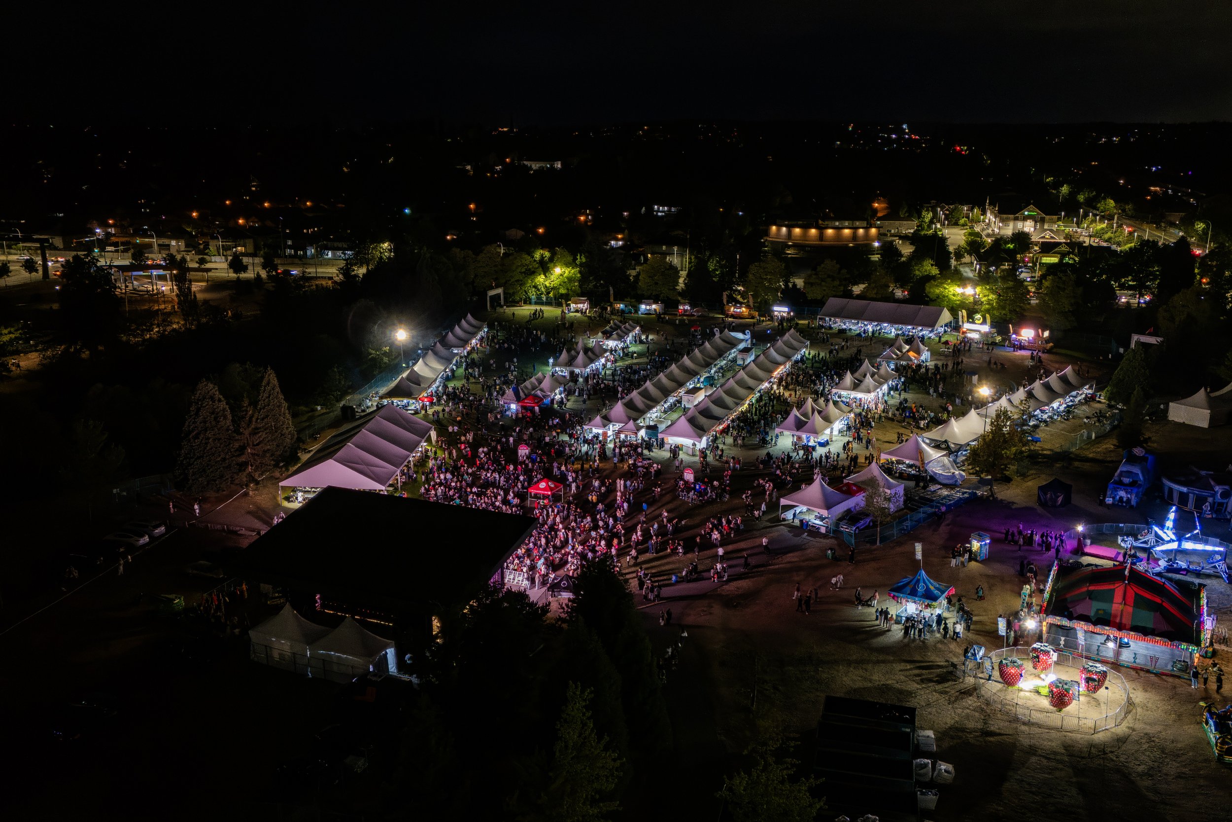 A night scene of an outdoor fair or carnival with multiple lit tents and a crowd of people.