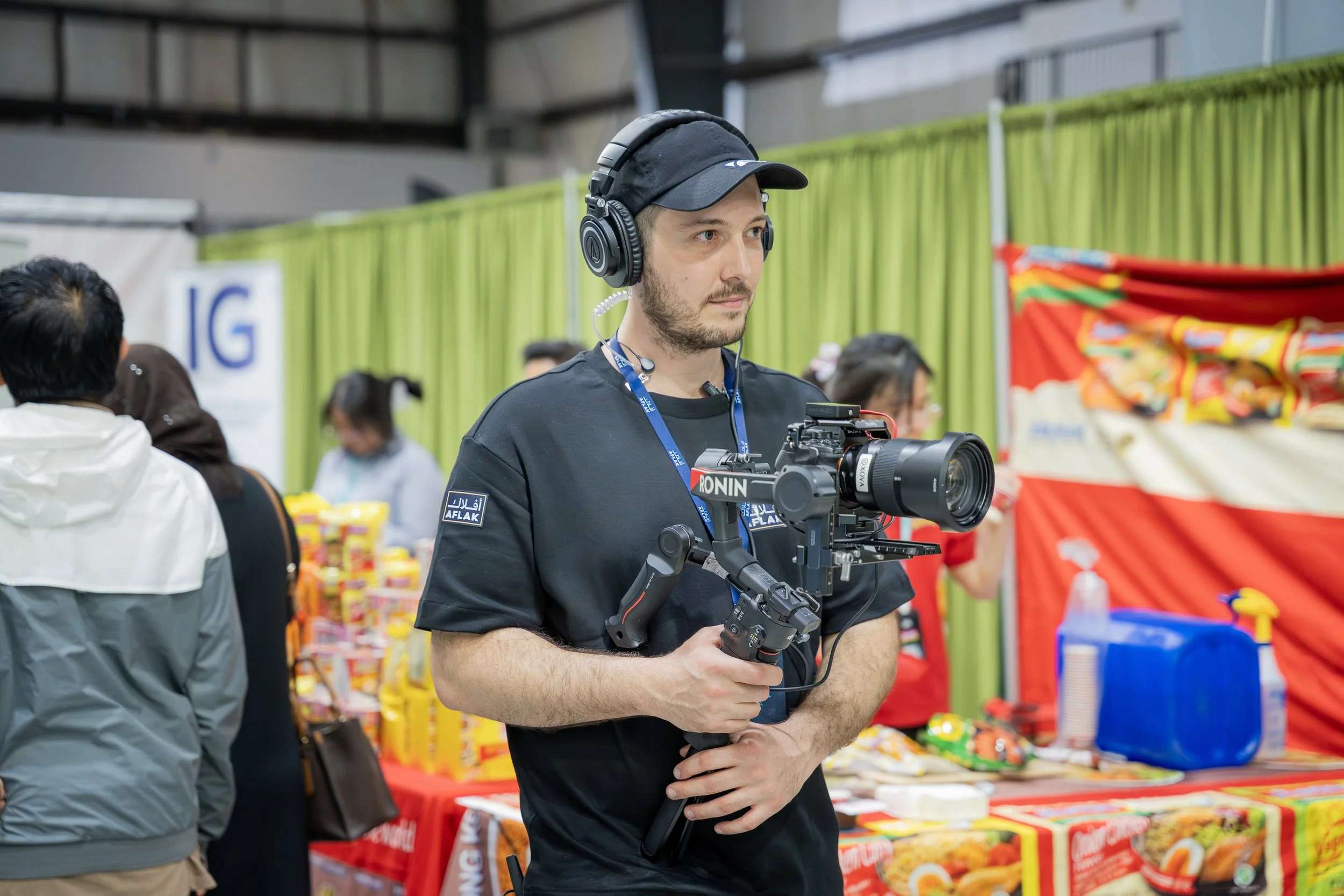 A man wearing a black shirt with 'AFLAK' patches on the sleeve, a black cap, and large headphones, holding a camera stabilizer at a public event. In the background, there are people and booths with colorful items and green curtains.