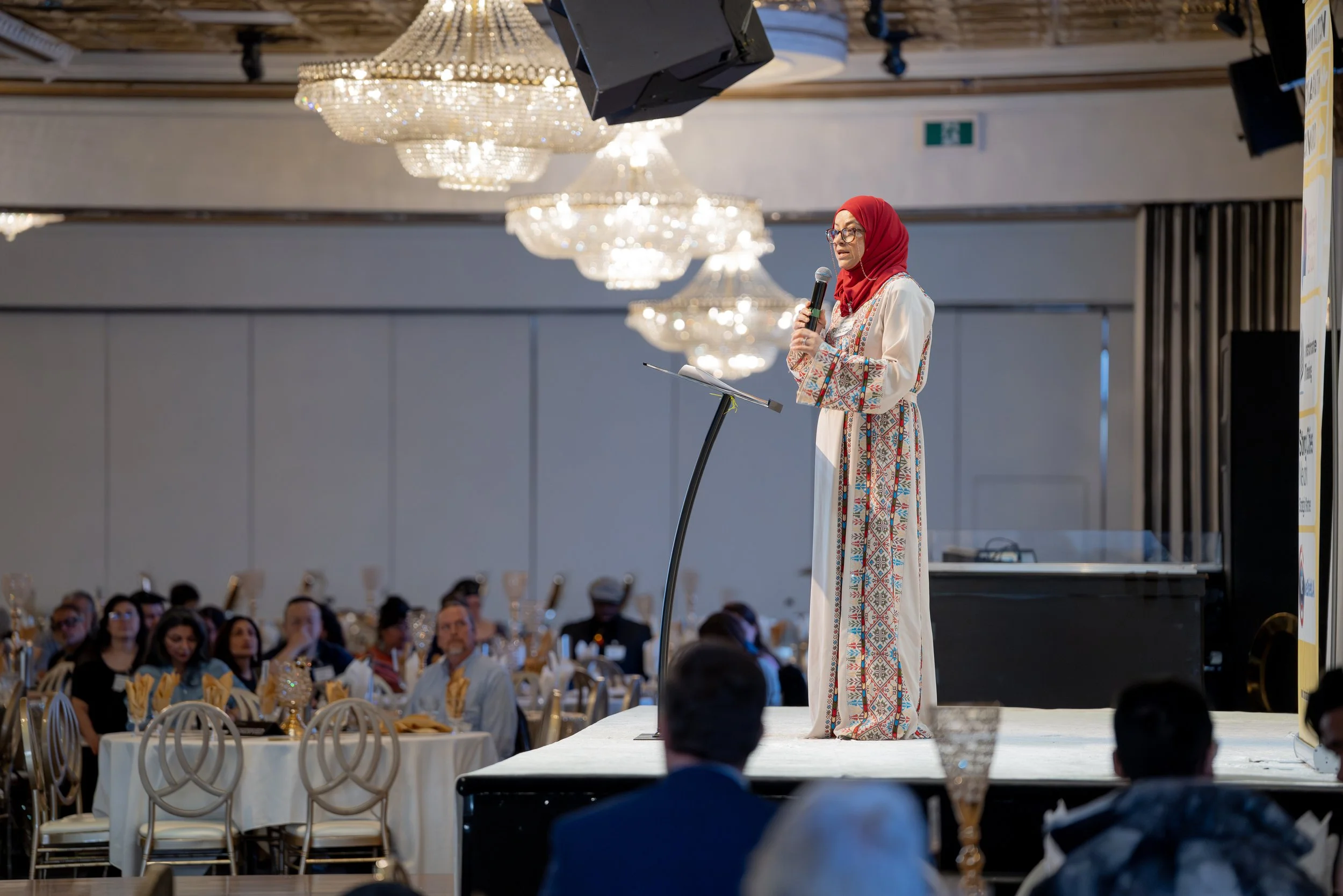 A woman in traditional attire is speaking at a podium during a conference or event in a large banquet hall, with chandeliers hanging from the ceiling and an audience seated at tables.