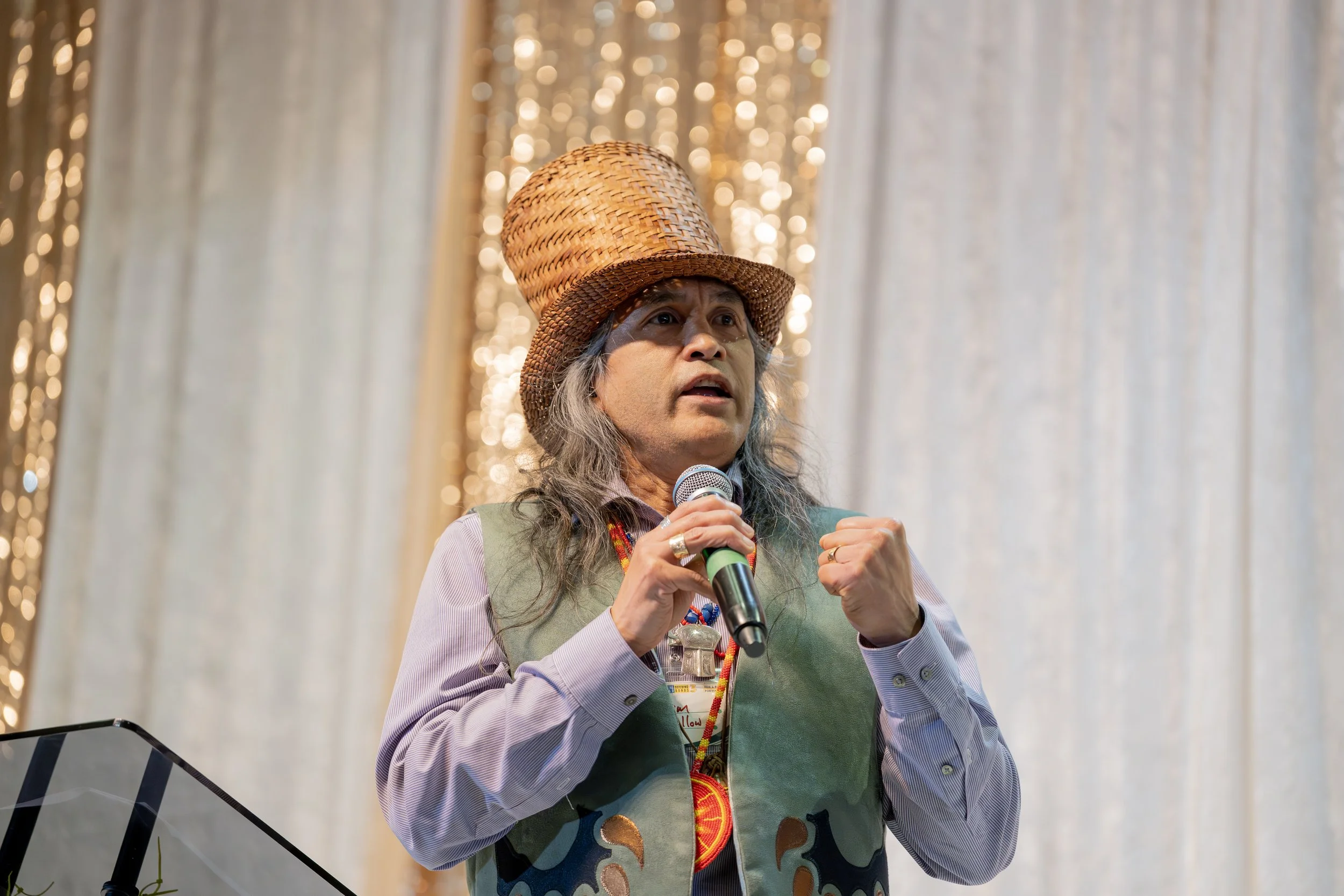 A person wearing a woven hat speaking into a microphone, standing in front of a curtain decorated with fairy lights.