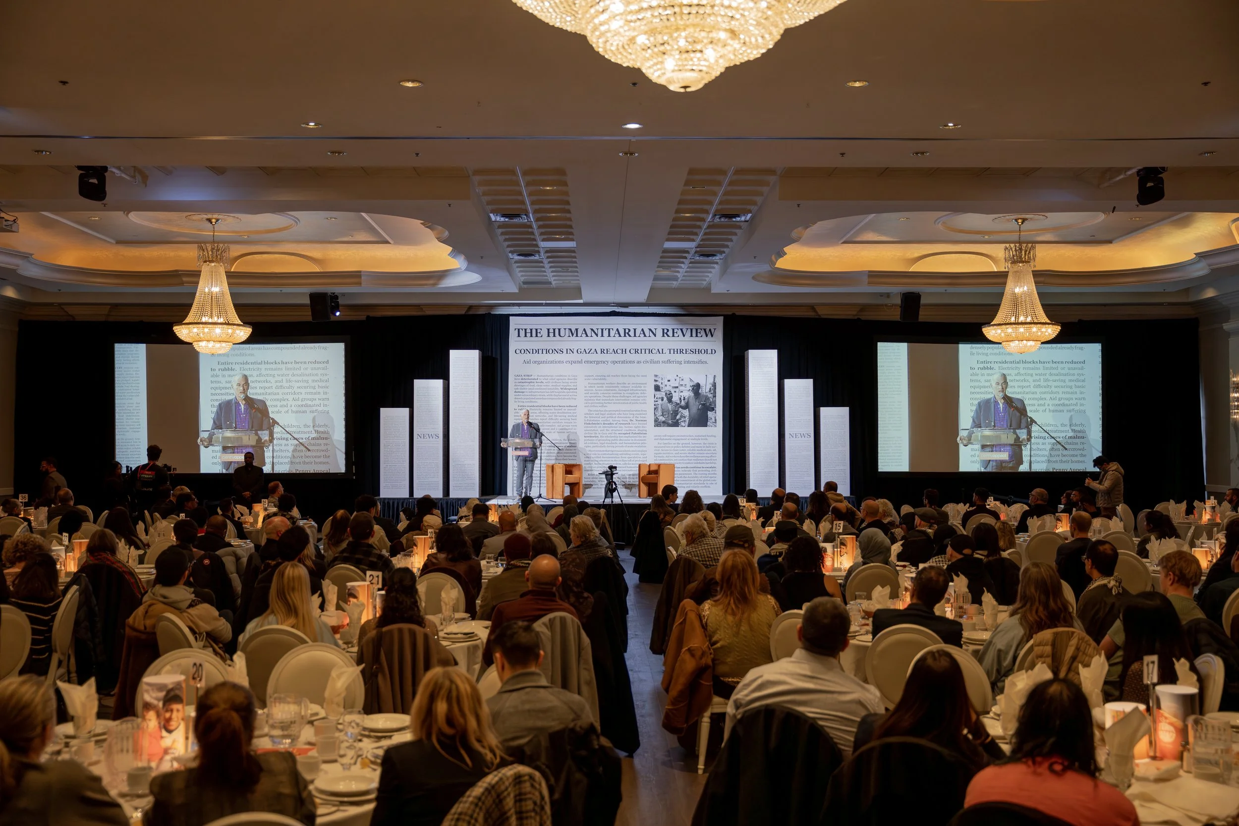 A large conference room filled with people sitting at round tables, facing a stage with a large banner titled "The Humanitarian Review" and a speaker standing at a podium. The room has ornate ceiling details and hanging chandeliers.