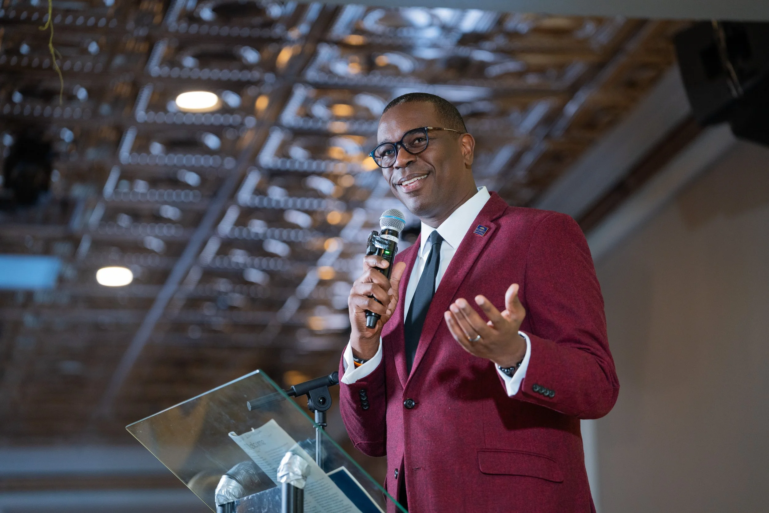 A man wearing glasses and a red blazer is speaking into a microphone while standing at a lectern with notes and a glass of water in front of him. The background features a wooden ceiling with lighting.