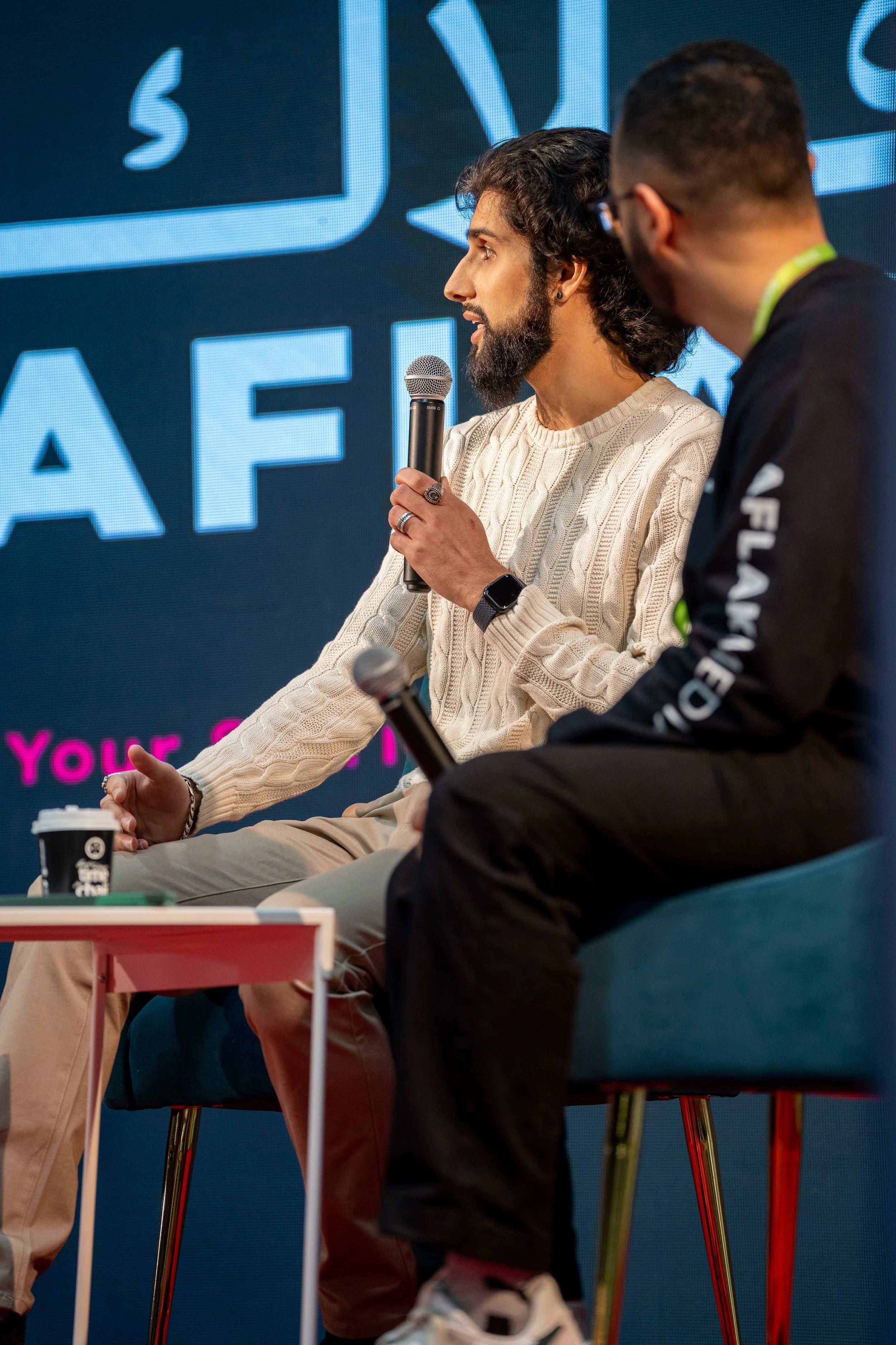 A man with dark curly hair, beard, and earrings is speaking into a microphone during an event, wearing a white cable-knit sweater. Another person with glasses and a black jacket is sitting nearby. Behind them is a large screen with partially visible text.