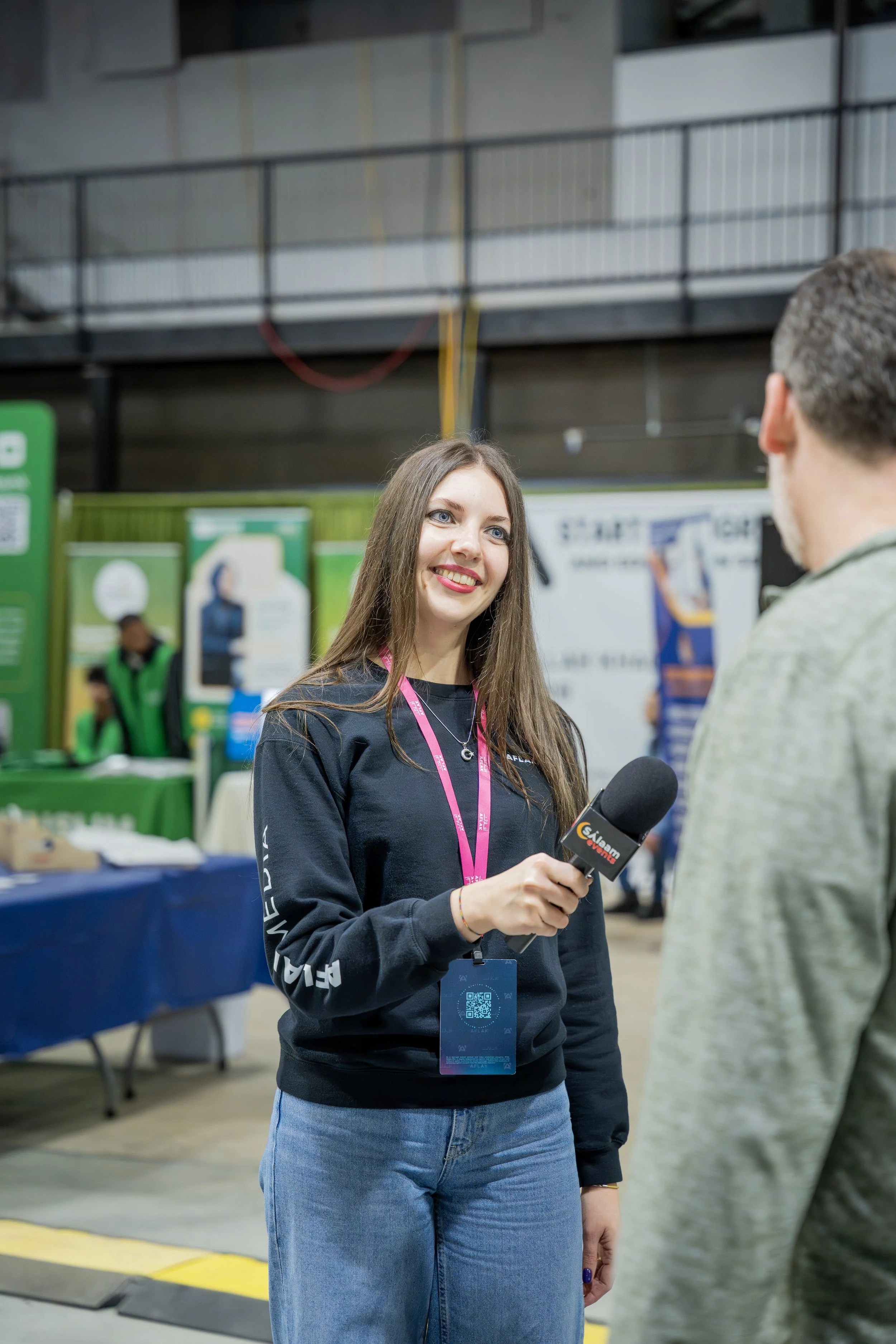 A young woman in a black long sleeve shirt holding a microphone, speaking to an older man at an indoor event or conference, with booths and banners in the background.