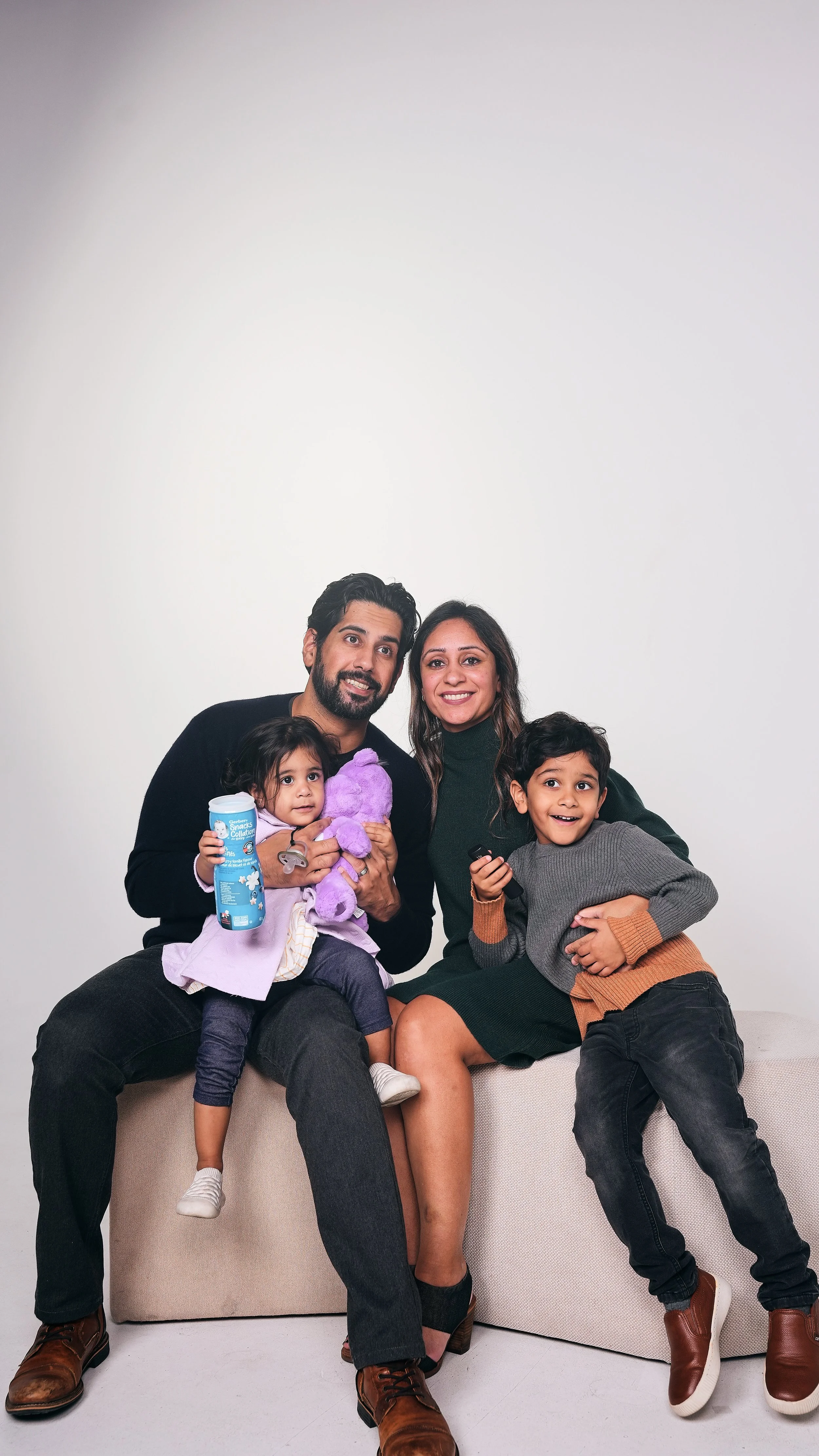 A family of four sitting on a beige couch against a plain white background. The father, with dark hair and a beard, is holding a young girl with dark hair, a pink dress, and a purple stuffed toy. The mother, with long dark hair, is smiling and sitting next to the father. A young boy with dark hair and a gray and orange sweater is leaning against her, smiling and holding a TV remote.