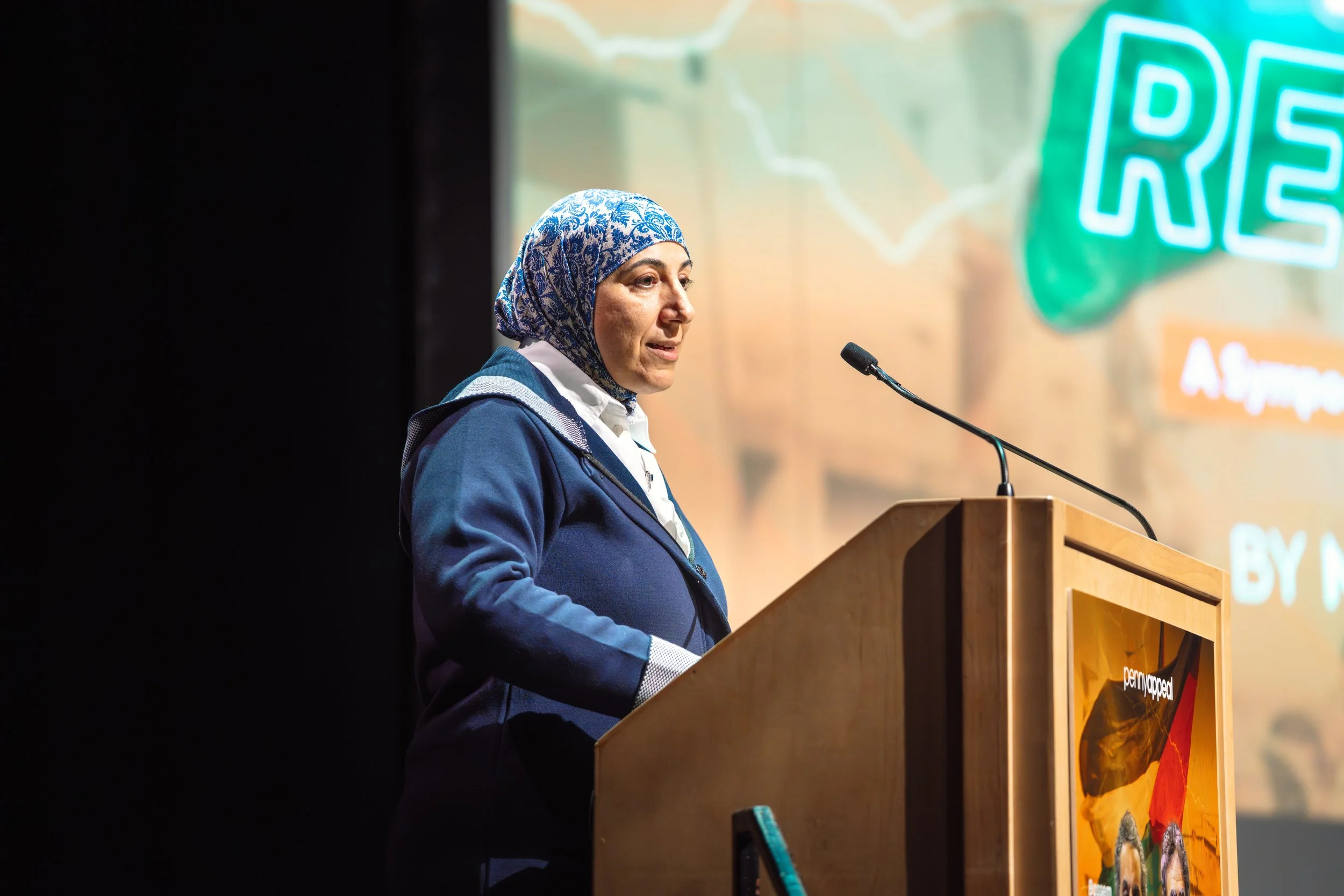 Woman wearing a patterned headscarf and blue blazer speaking at a podium during an event with a colorful backdrop featuring the word 'RES' in neon letters.