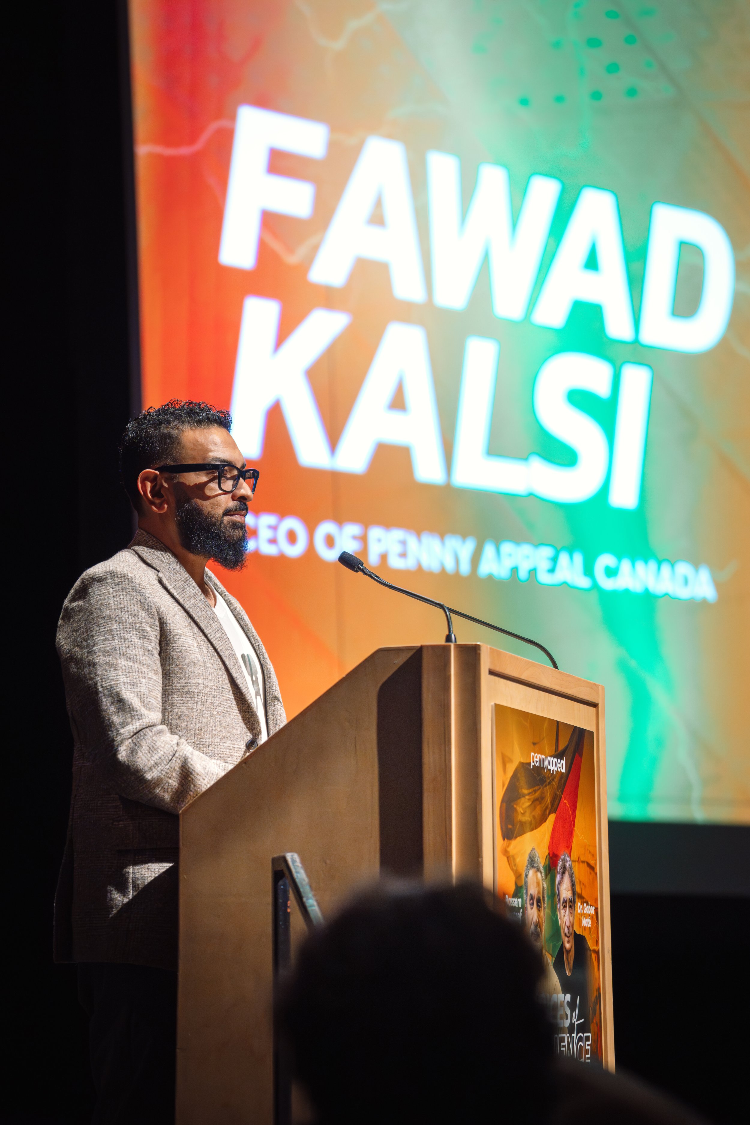 A man with glasses and a beard in a brown blazer is standing at a wooden podium, speaking into a microphone. Behind him is a large, colorful screen with text that reads 'FAWAD KALSI' and additional smaller text about a PENNY APPEAL CANADA event. The background of the screen includes a vibrant, abstract design.