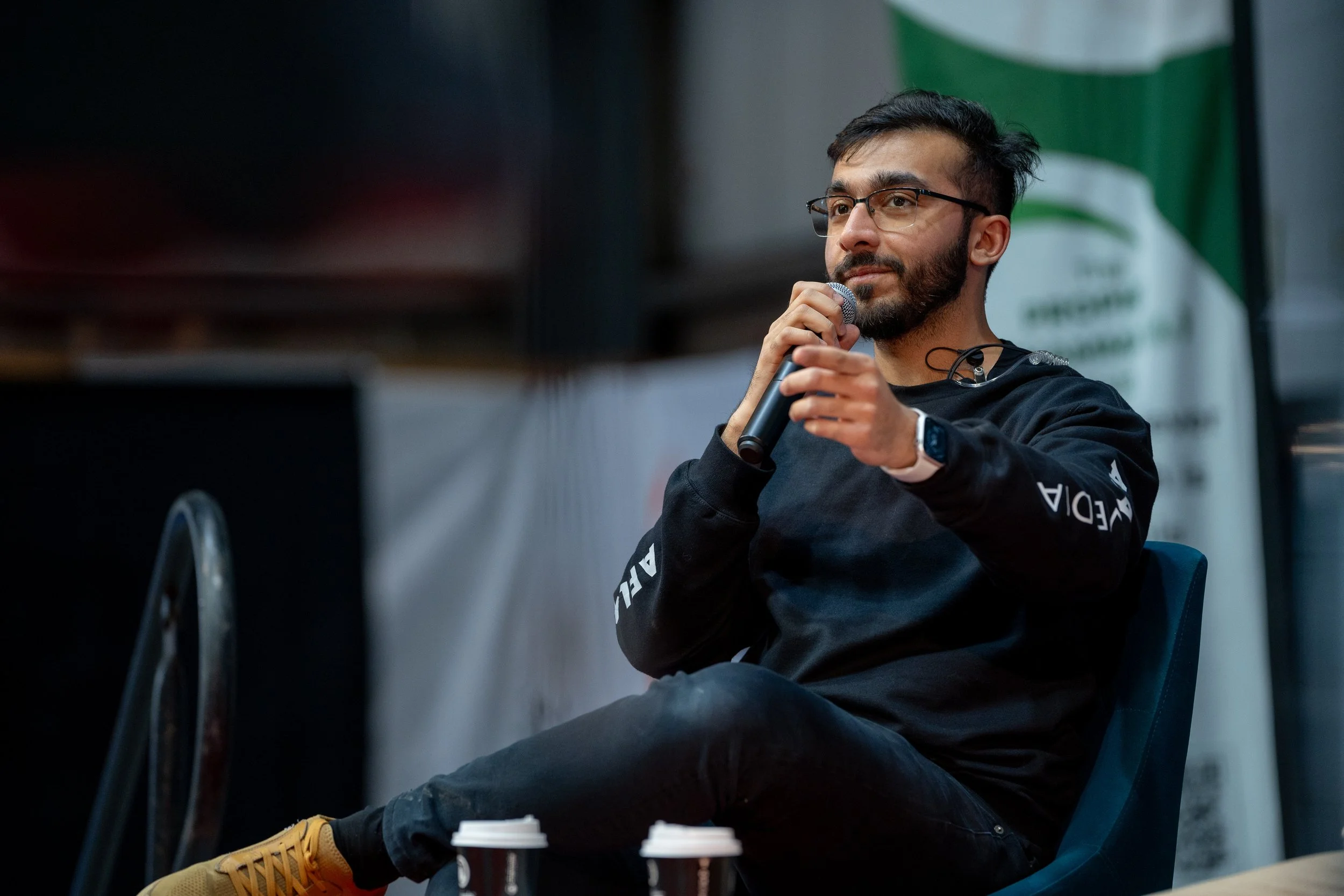 A man with glasses, beard, and dark hair sitting outdoors, holding a microphone near his mouth, wearing a black hoodie with white text, with coffee cups on the bench in front of him, and a green and white flag in the background.