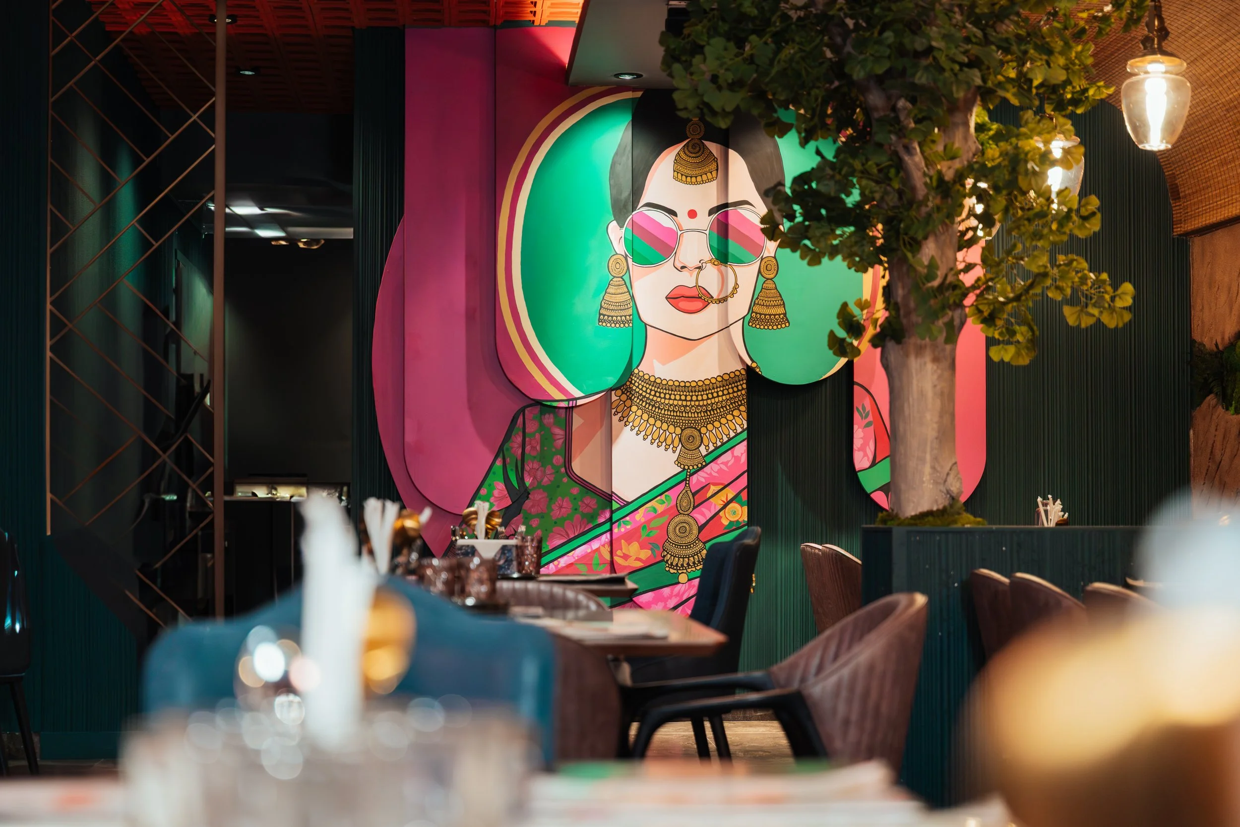 Colorful mural of a woman wearing sunglasses, traditional jewelry, and a sari, with a green and pink background, inside a restaurant or cafe.