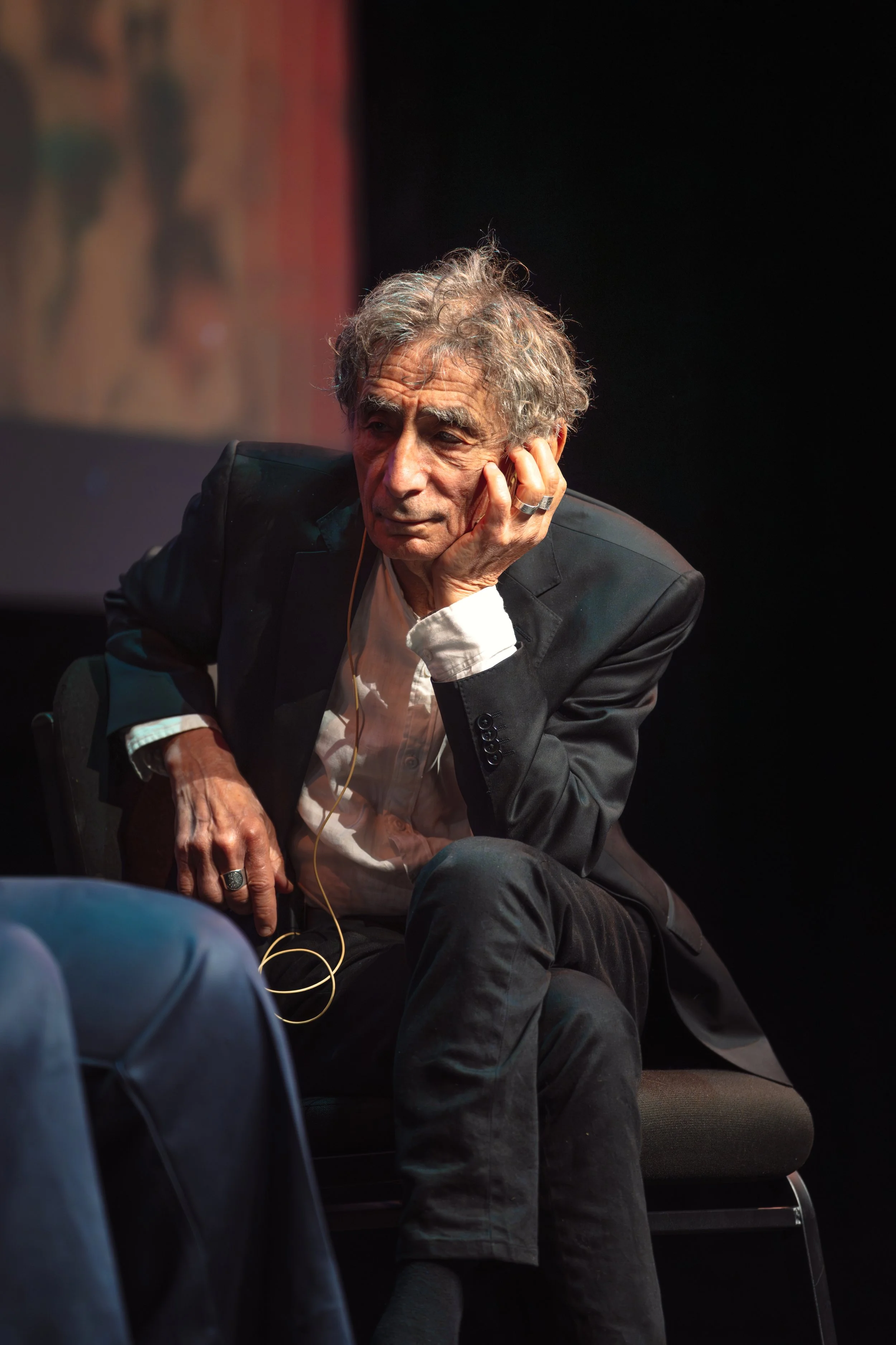 An elderly man with grey, curly hair and a serious expression, sitting and resting his head on his hand, wearing a black blazer and white shirt, in a dark room with a blank background.