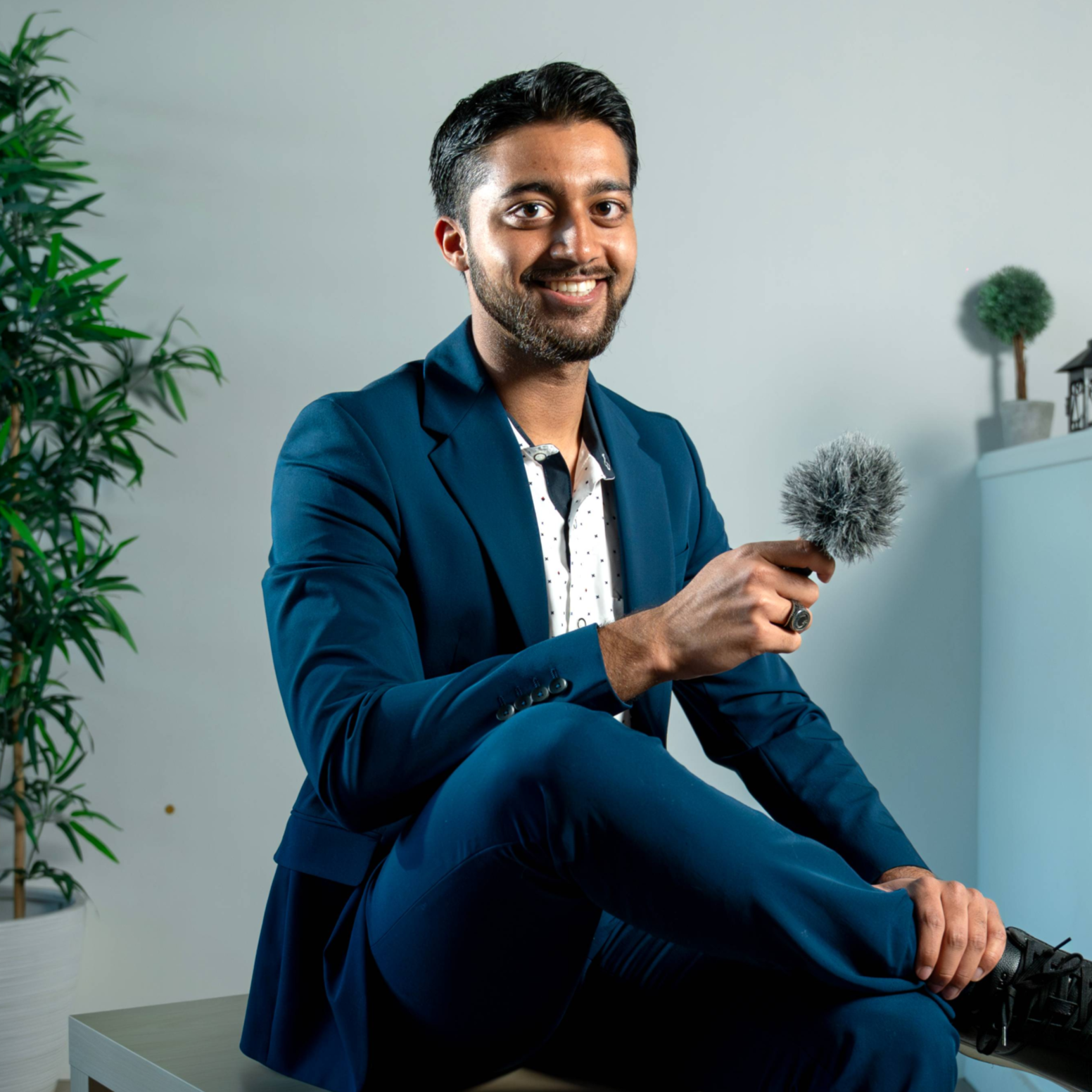 A smiling man in a navy suit sitting indoors, holding a microphone with a windscreen, with a plant and decorative items in the background.