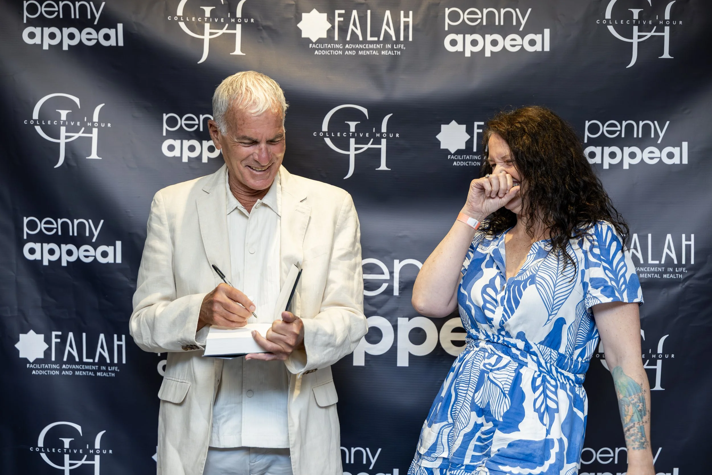 Two people smiling and laughing during a signing event with a dark backdrop featuring logos for Penny Appeal, Falah, and Collective Hour.