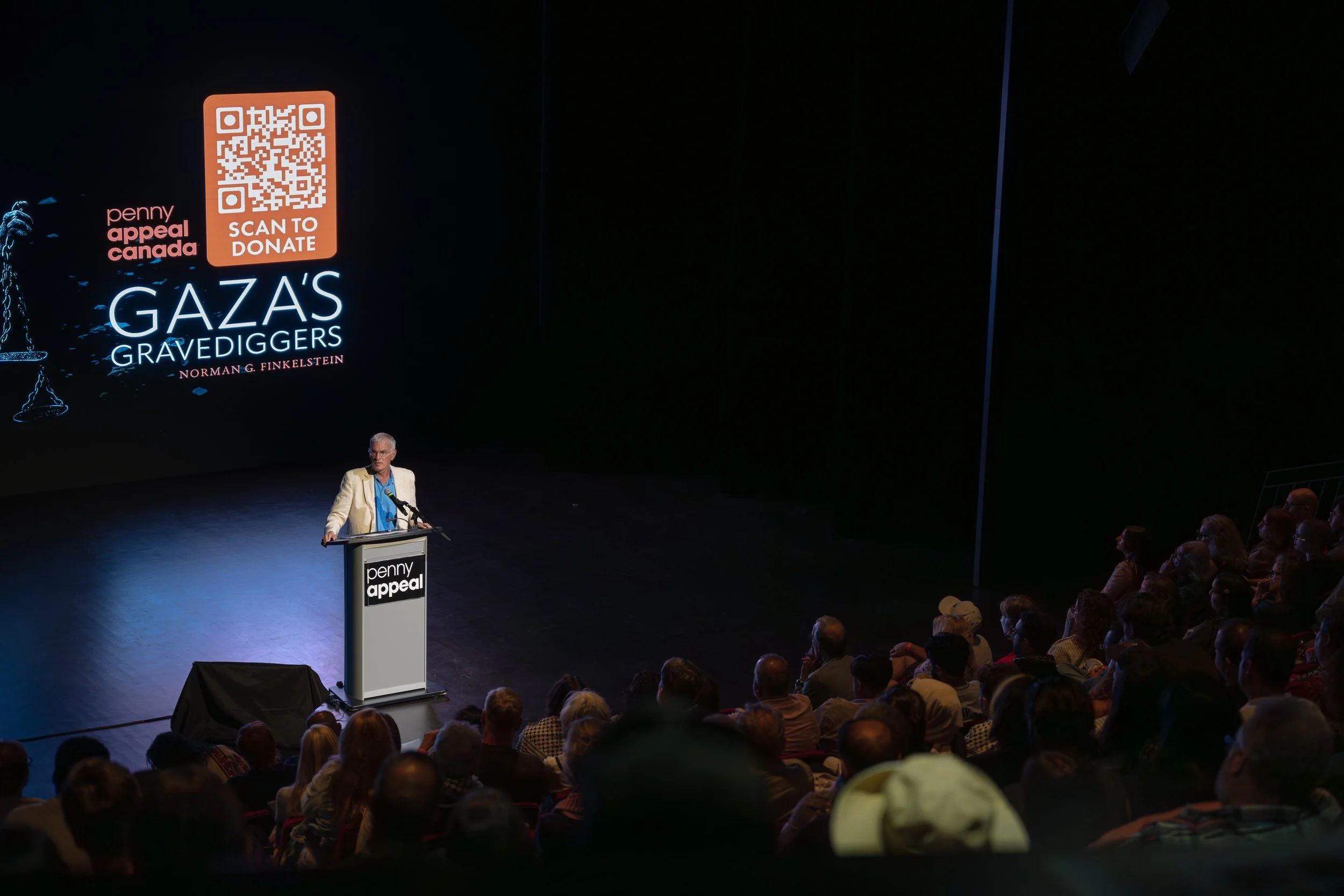A speaker is standing at a podium on a stage, addressing an audience for a Pennies Appeal Canada event. The background screen displays logos, a QR code with 'SCAN TO DONATE,' and text about Gaza's grave diggers by Norman G. Finkelstein. There are people sitting and listening attentively in a dark conference hall.
