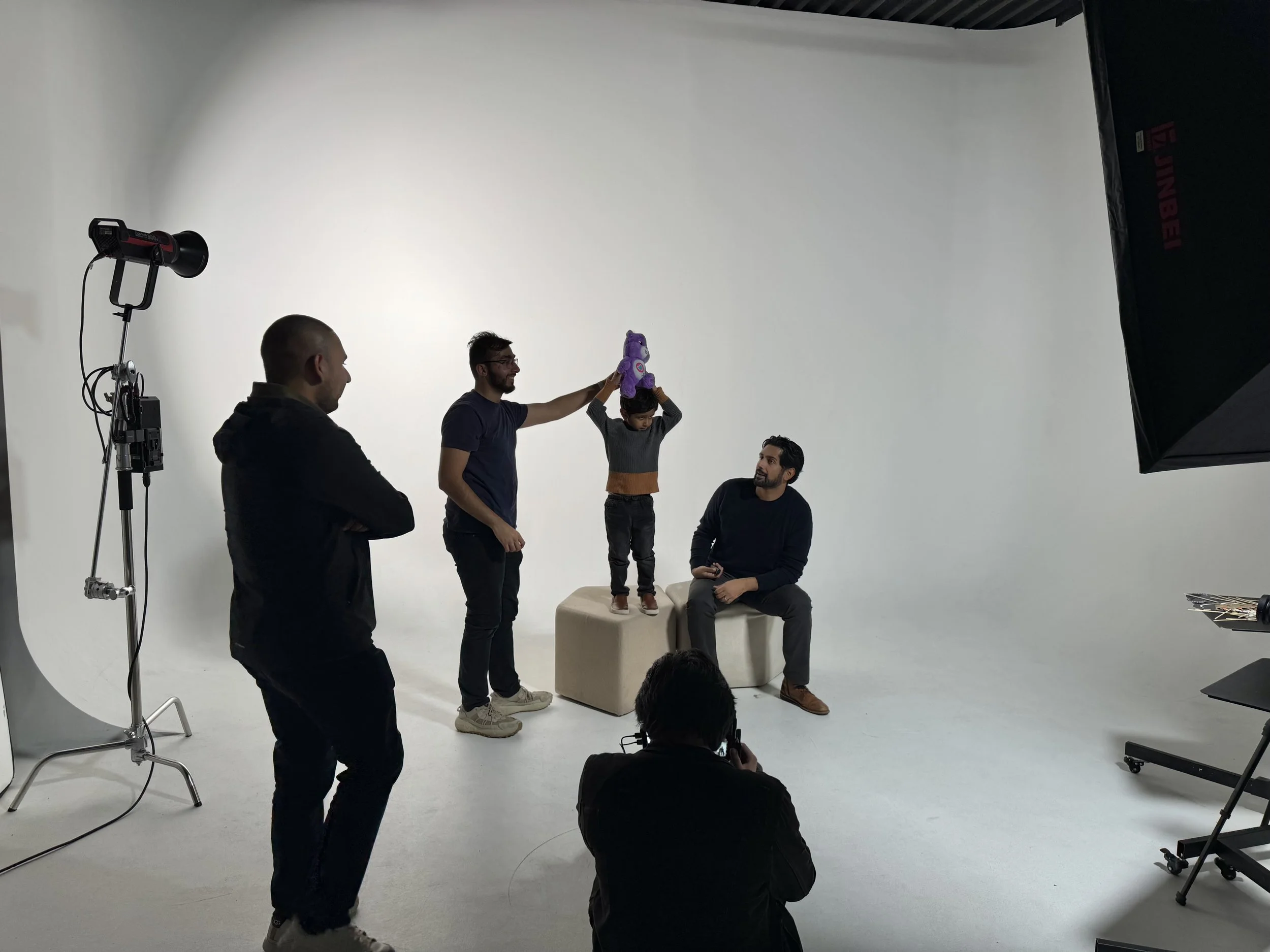 People in a photo studio setting, with some engaged in a photo session involving a child holding a purple stuffed animal on an adult's shoulders, while others observe or work with photography equipment, including lighting and a plain white background.