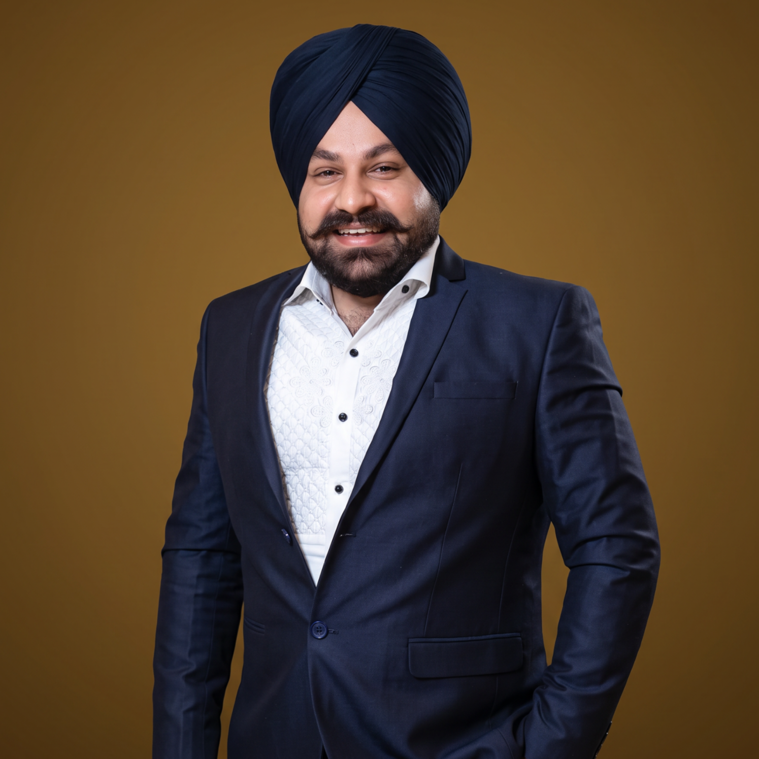 A man wearing a blue turban and a navy blue suit with a white shirt, smiling against a brown background.