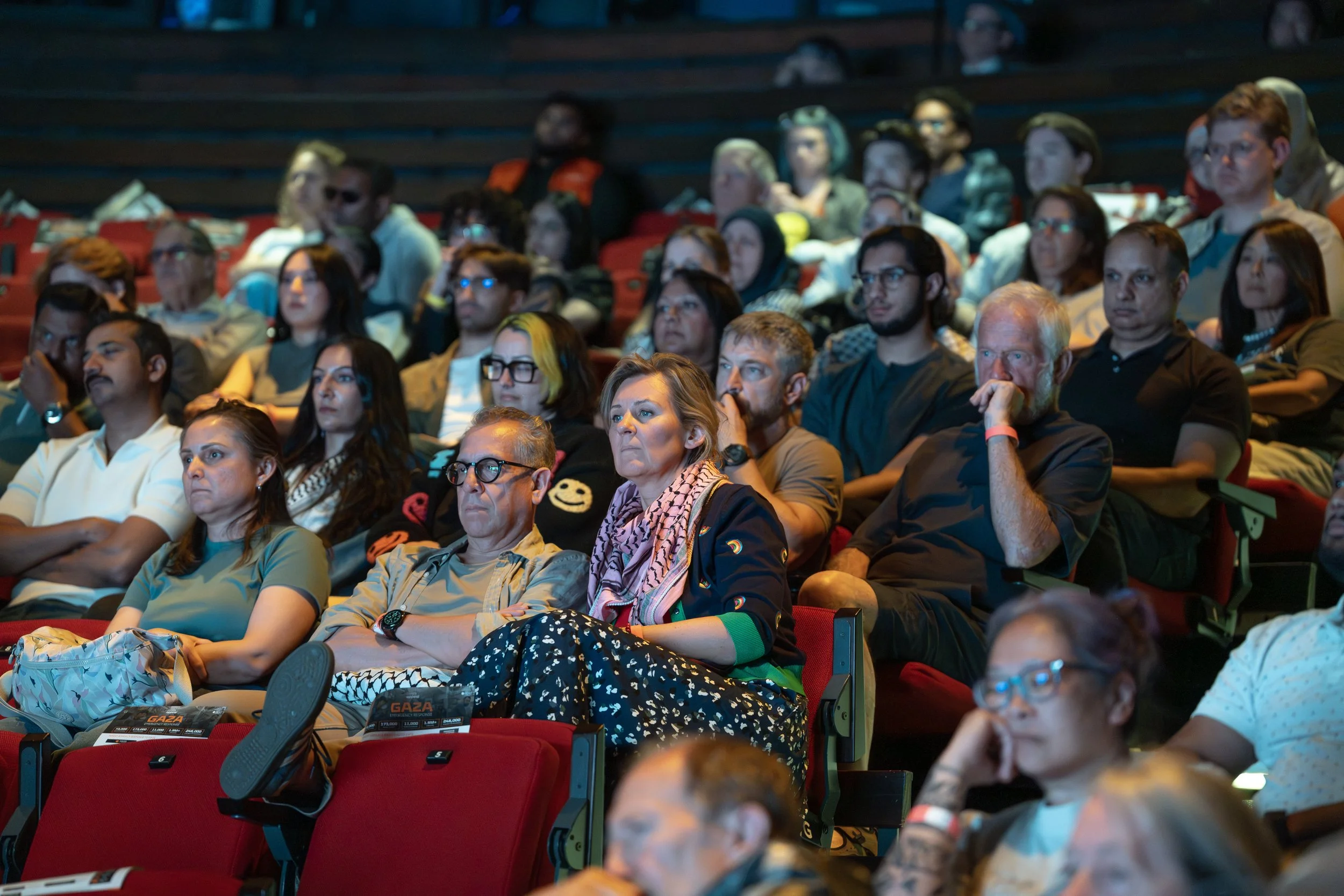 Audience watching a presentation or event in a theater or auditorium with red seats and dim lighting.