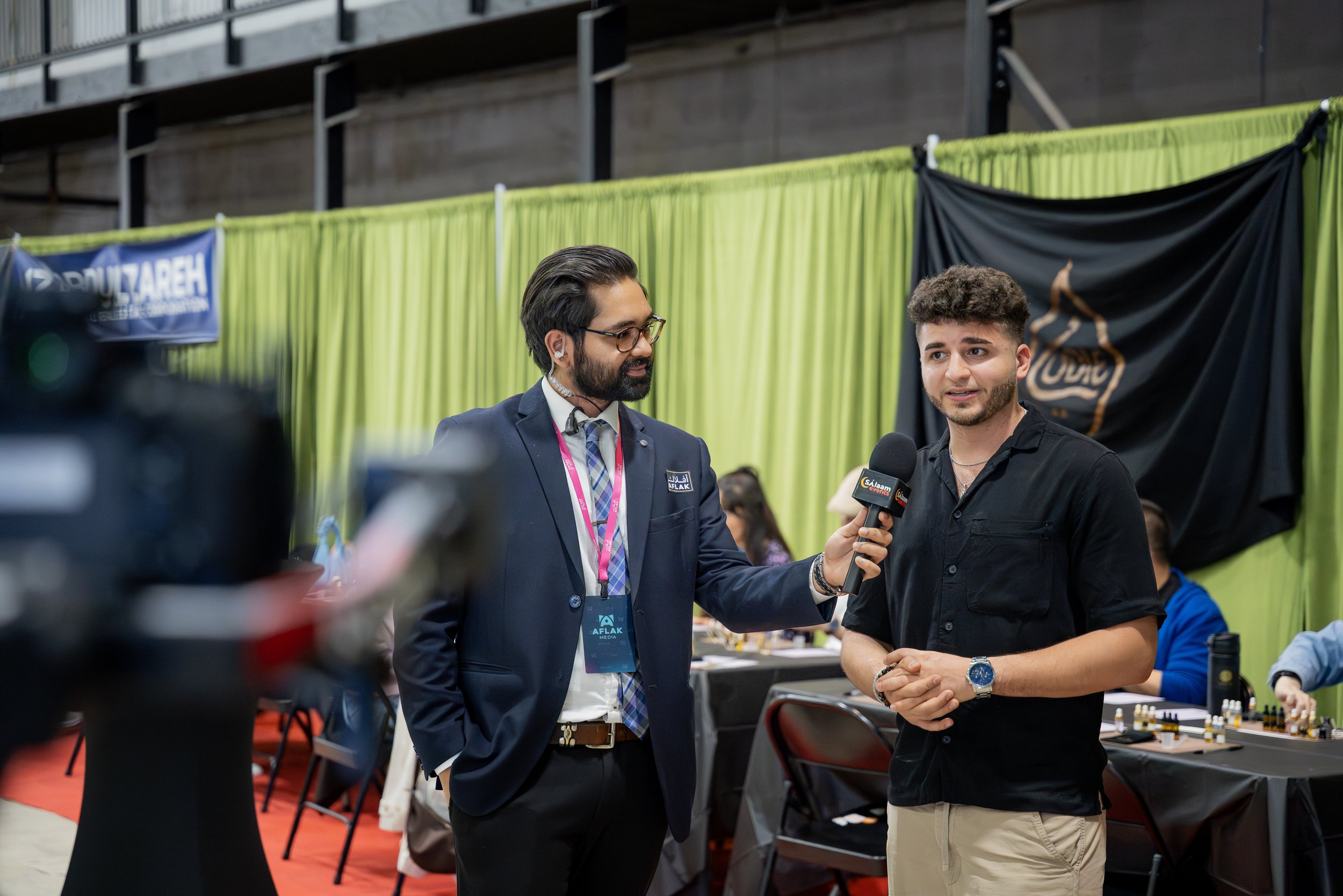 Two men are engaged in an interview at a conference or event. One man, in a suit and glasses, is holding a microphone and talking to the other man, who is dressed in a black shirt and khaki pants. There is a green and black backdrop behind them with some tables and chairs nearby.