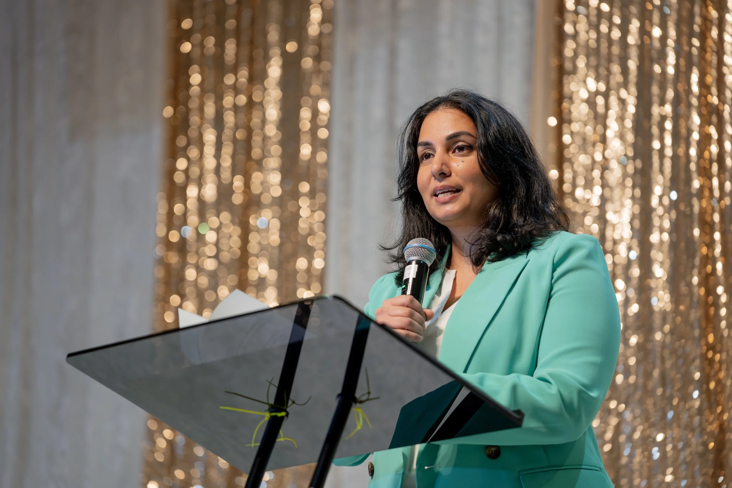 Woman wearing a turquoise blazer speaking into a microphone at an event with a decorated background of hanging shiny gold beads.