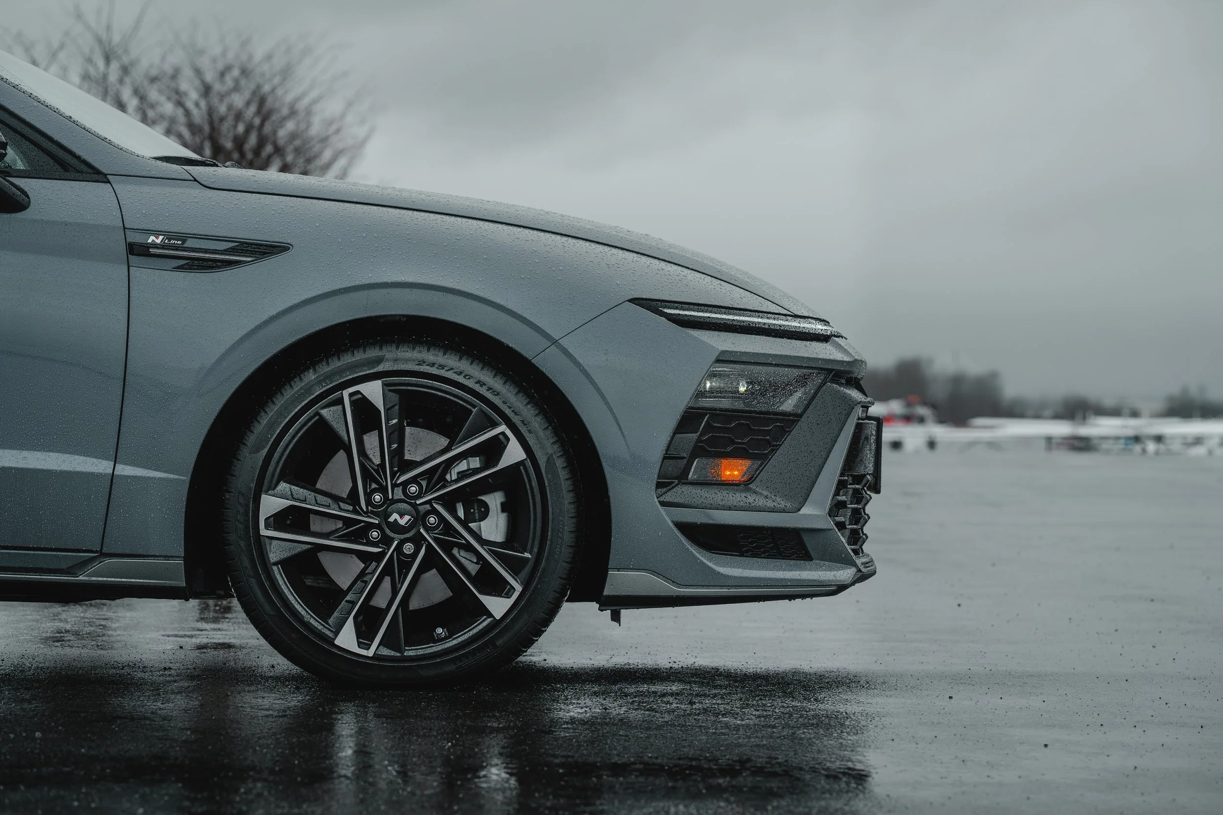 Close-up of the front part of a gray modern sports car with black rims, parked on a wet surface on a cloudy day. The background shows a blurry winter landscape with leafless trees and snow.